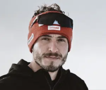 Man wearing a red headband, black sunglasses pushed up on his head, and a black hoodie, against a plain background.