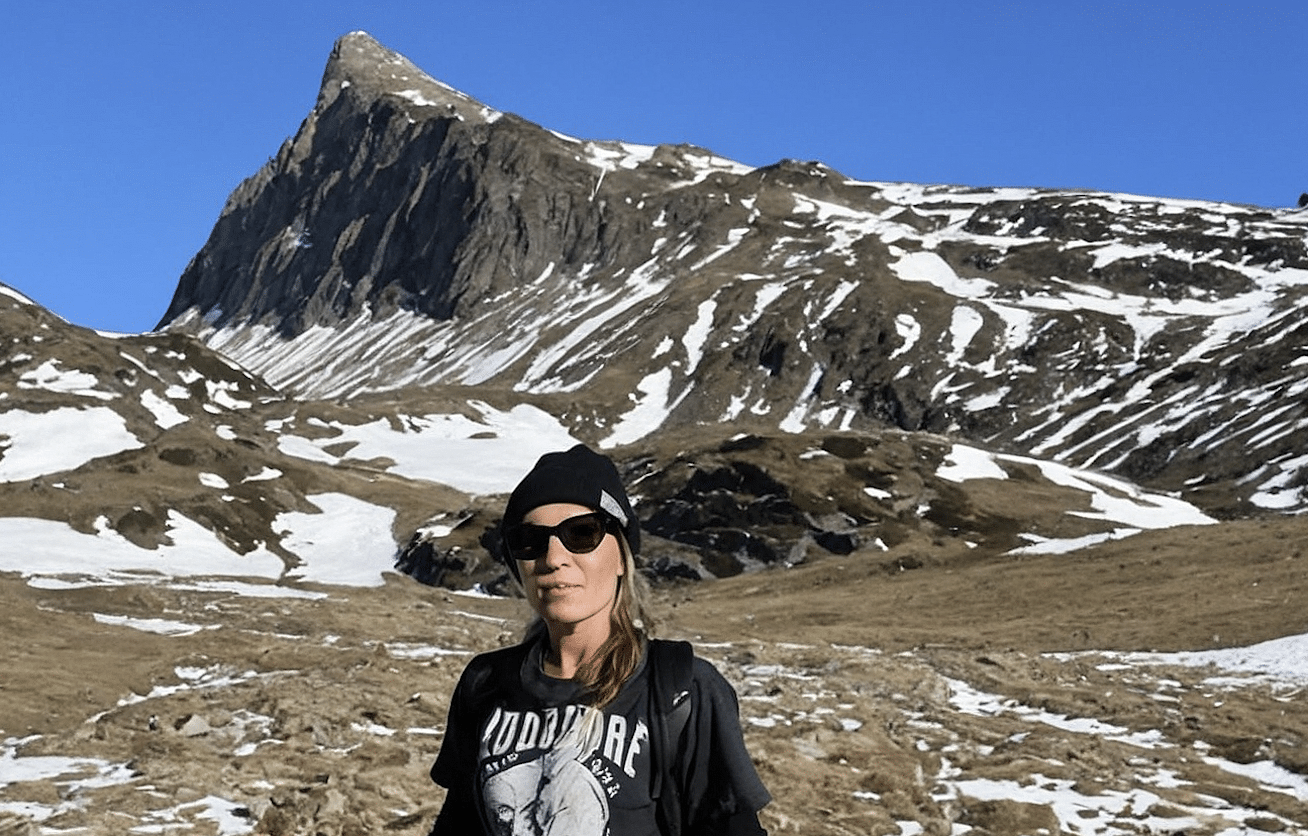 Woman wearing sunglasses, a black beanie, and a black T-shirt with a graphic design, standing in a mountainous landscape with patches of snow and a prominent rocky mountain peak in the background.