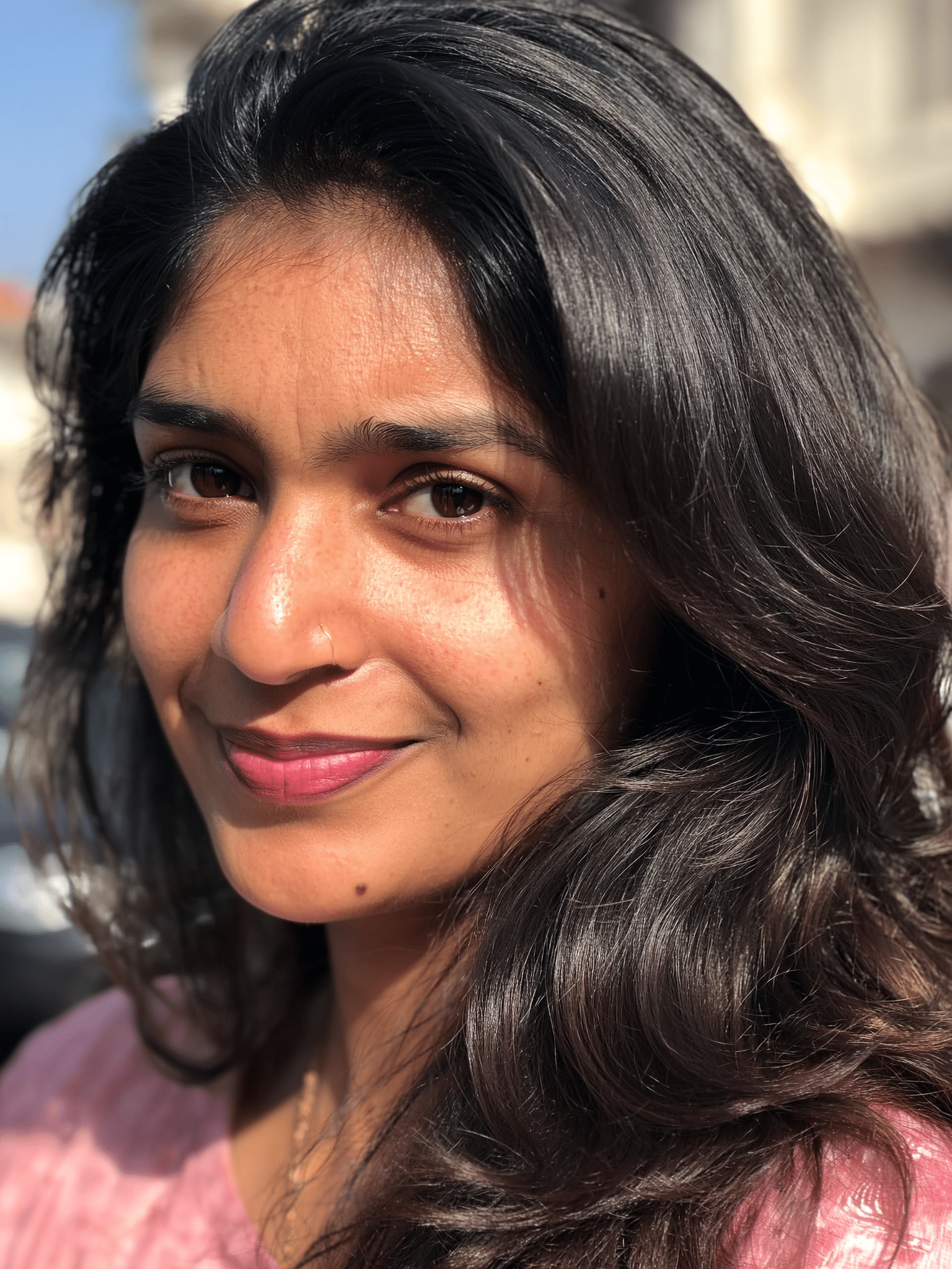 Close-up of a woman with dark hair smiling outdoors in sunlight, wearing a pink top.