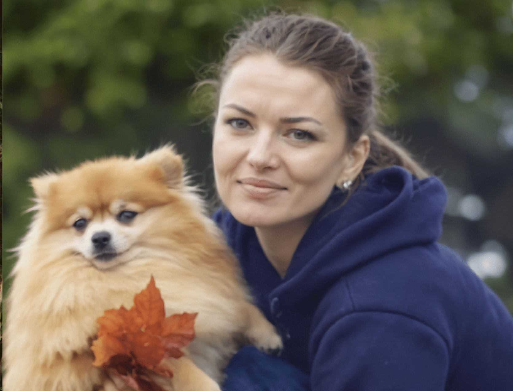 A woman in a blue hoodie holding a small fluffy Pomeranian dog with a autumn leaf in its fur outdoors, with blurred green trees in the background.