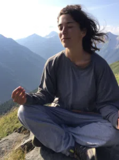 A woman sitting cross-legged outdoors on a mountain overlooking a scenic valley with mountains and blue sky.