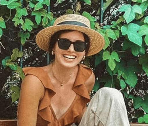 Woman with large black sunglasses and a straw hat, sitting outdoors near green leafy plants, smiling.