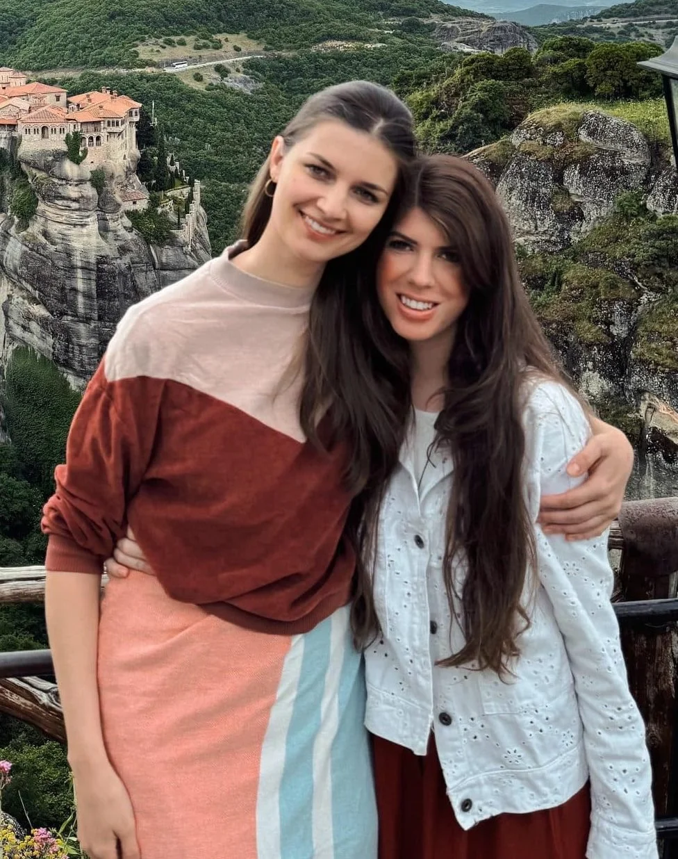 Two young women smiling and embracing each other outdoors with a mountainous landscape and buildings on cliffs in the background.