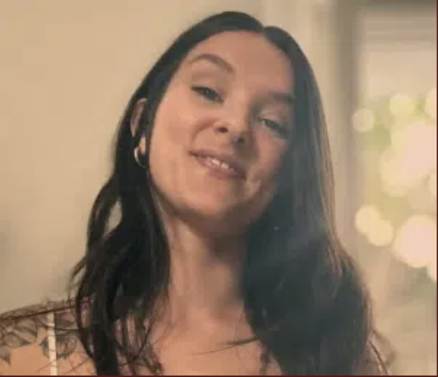 A woman with long dark hair, smiling with a slight tilt to her head, wearing earrings, indoors with bokeh lights in the background.