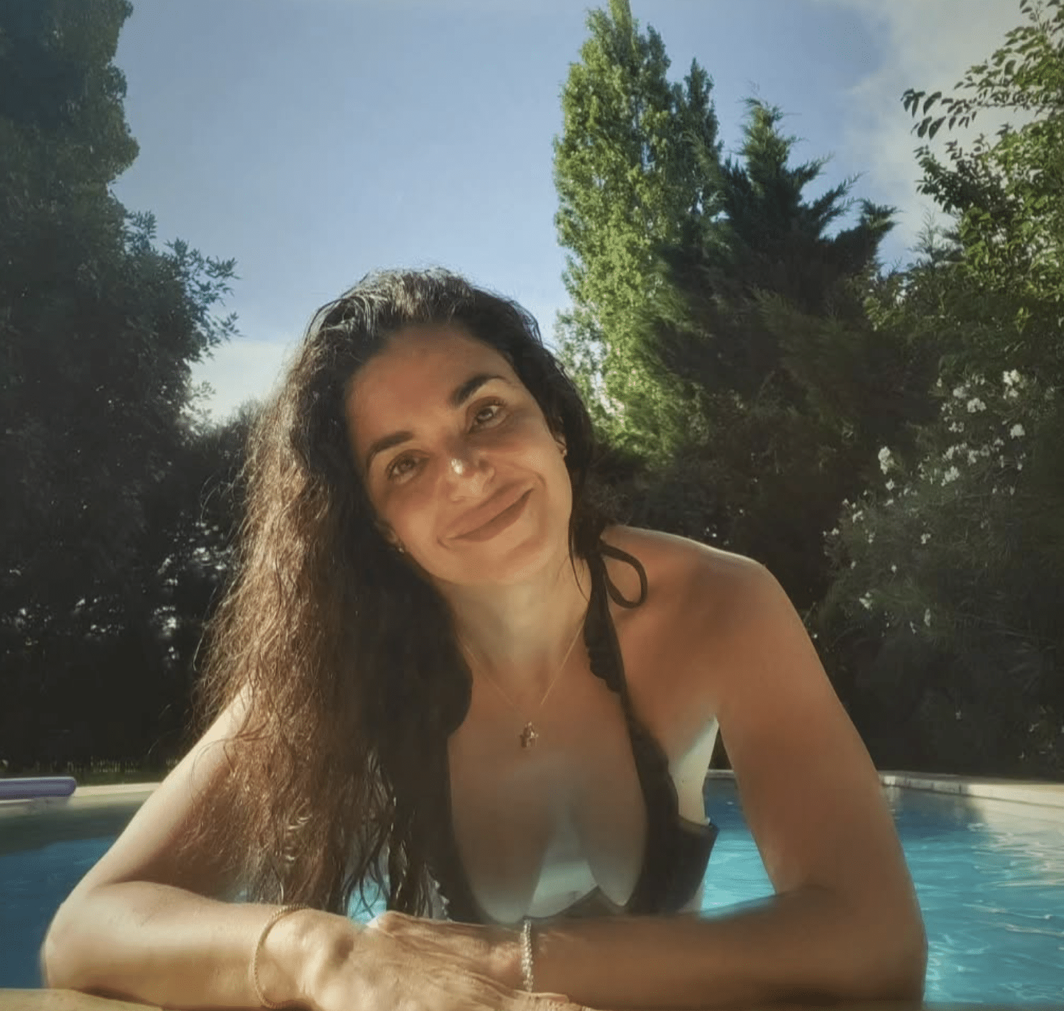 Smiling woman with wet hair leaning on pool edge outdoors, with tall green trees and a blue sky in the background.