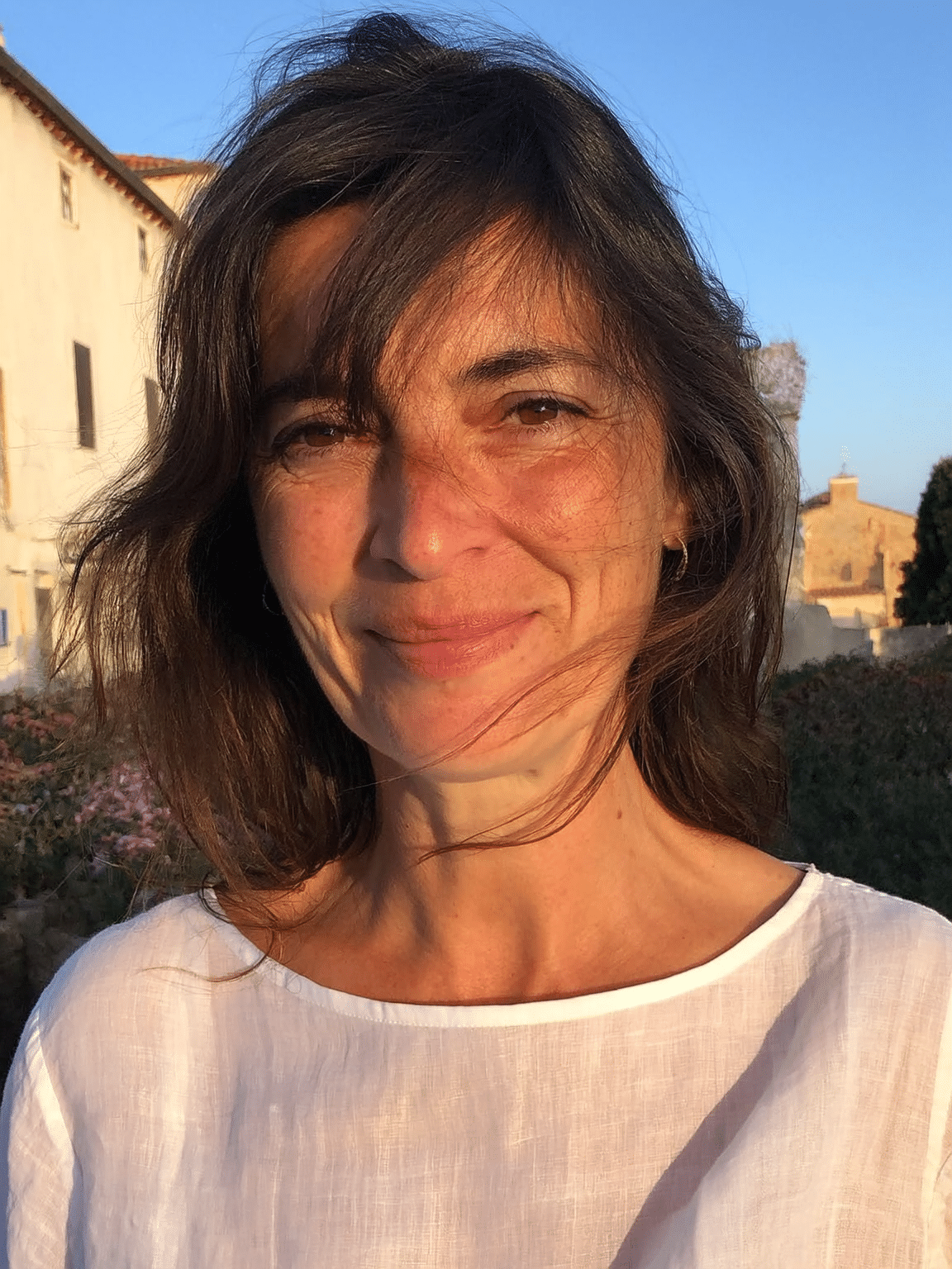 A woman with shoulder-length dark hair, smiling, outdoors during sunset, wearing a white top with a round neckline, with buildings and clear blue sky in the background.