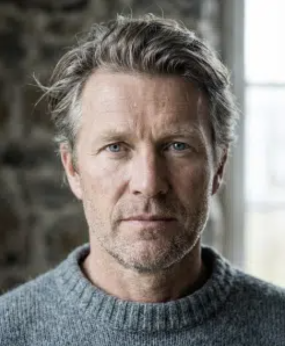 Close-up of a middle-aged man with gray hair and blue eyes, wearing a gray sweater, standing indoors with a brick wall and window in the background.