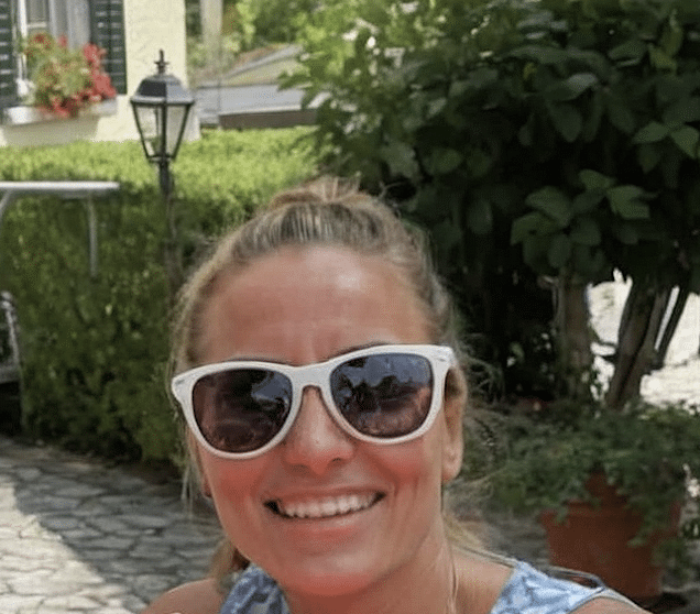 Woman smiling outdoors wearing large white sunglasses, with greenery and a lamp post in the background.