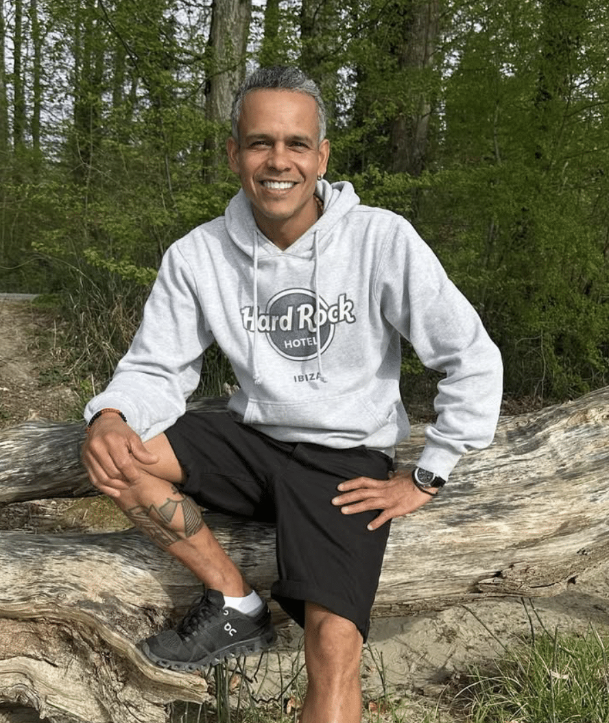 A man smiling and sitting on a large fallen tree trunk in a forest with green foliage in the background. He is wearing a gray Hard Rock Hotel Ibiza hoodie, black shorts, and black athletic shoes, with one hand resting on his knee and the other on the log.