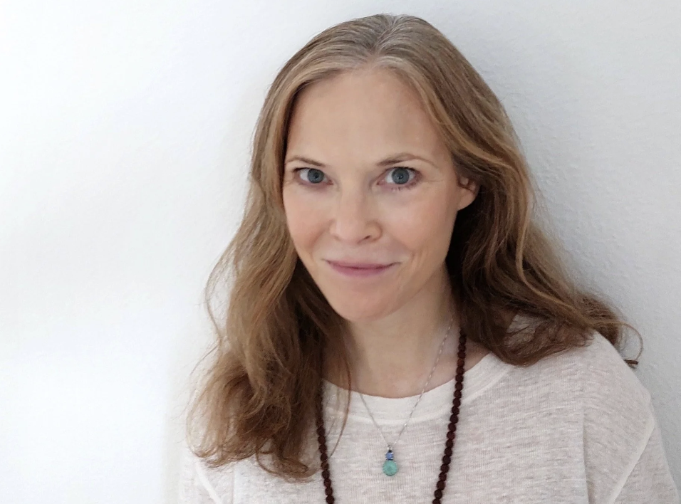 A woman with long, wavy red hair and blue eyes smiling softly in front of a plain, white wall.