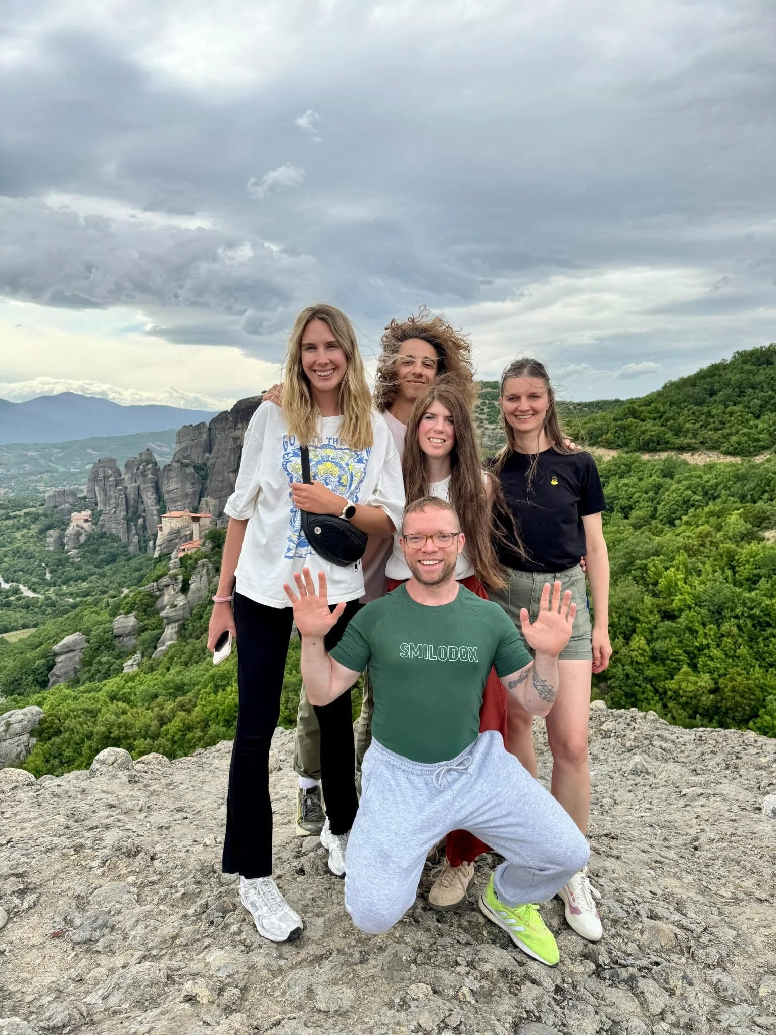 Group of six friends posing on a rocky hill with green hills and large rock formations in the background, under cloudy sky.