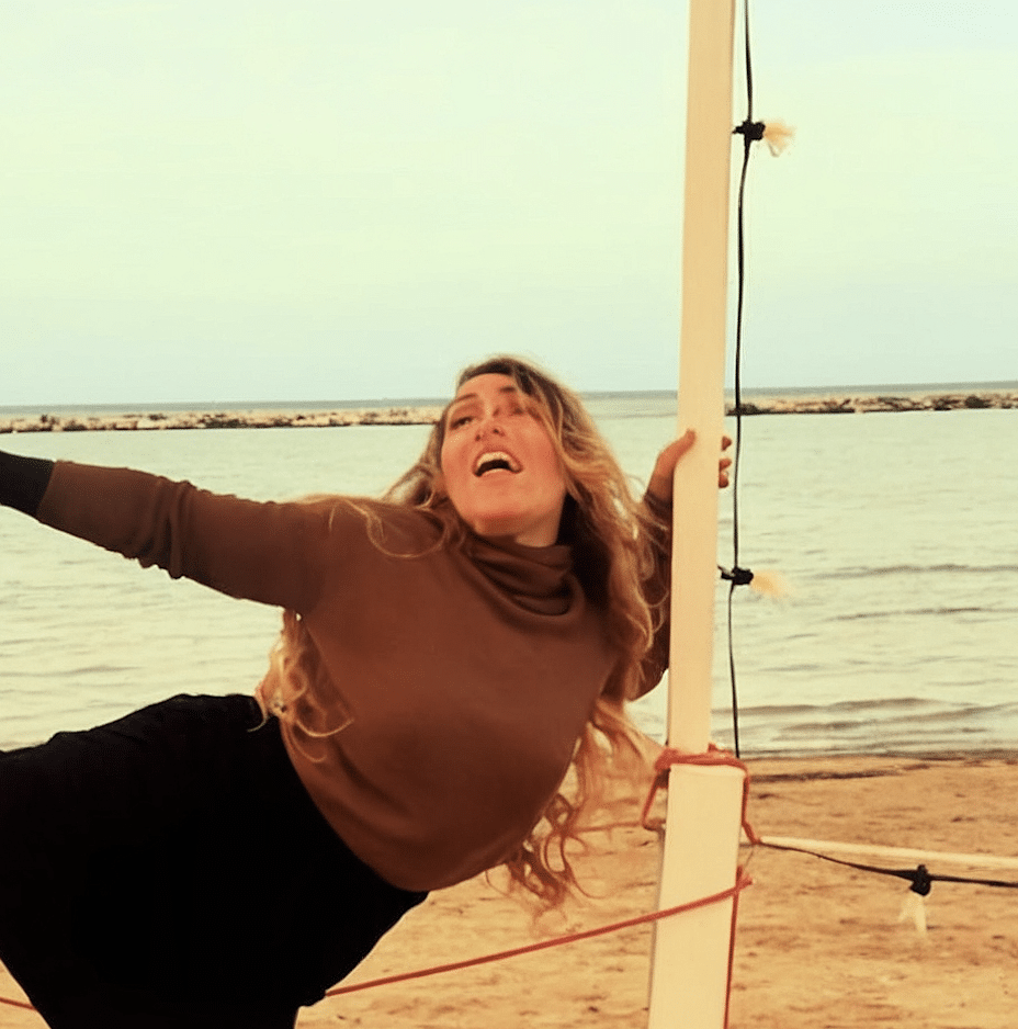 Woman in a brown long-sleeve top and black pants at the beach, holding onto a volleyball net pole, smiling with arms outstretched.