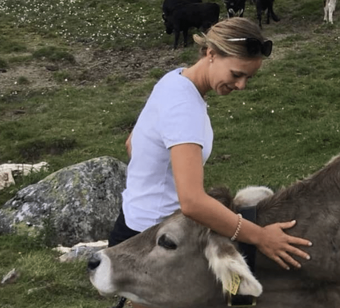 A woman interacting with a calf outdoors in a grassy area with rocks. The woman is smiling and touching the calf, which has a yellow ear tag. In the background, there are several cows grazing.