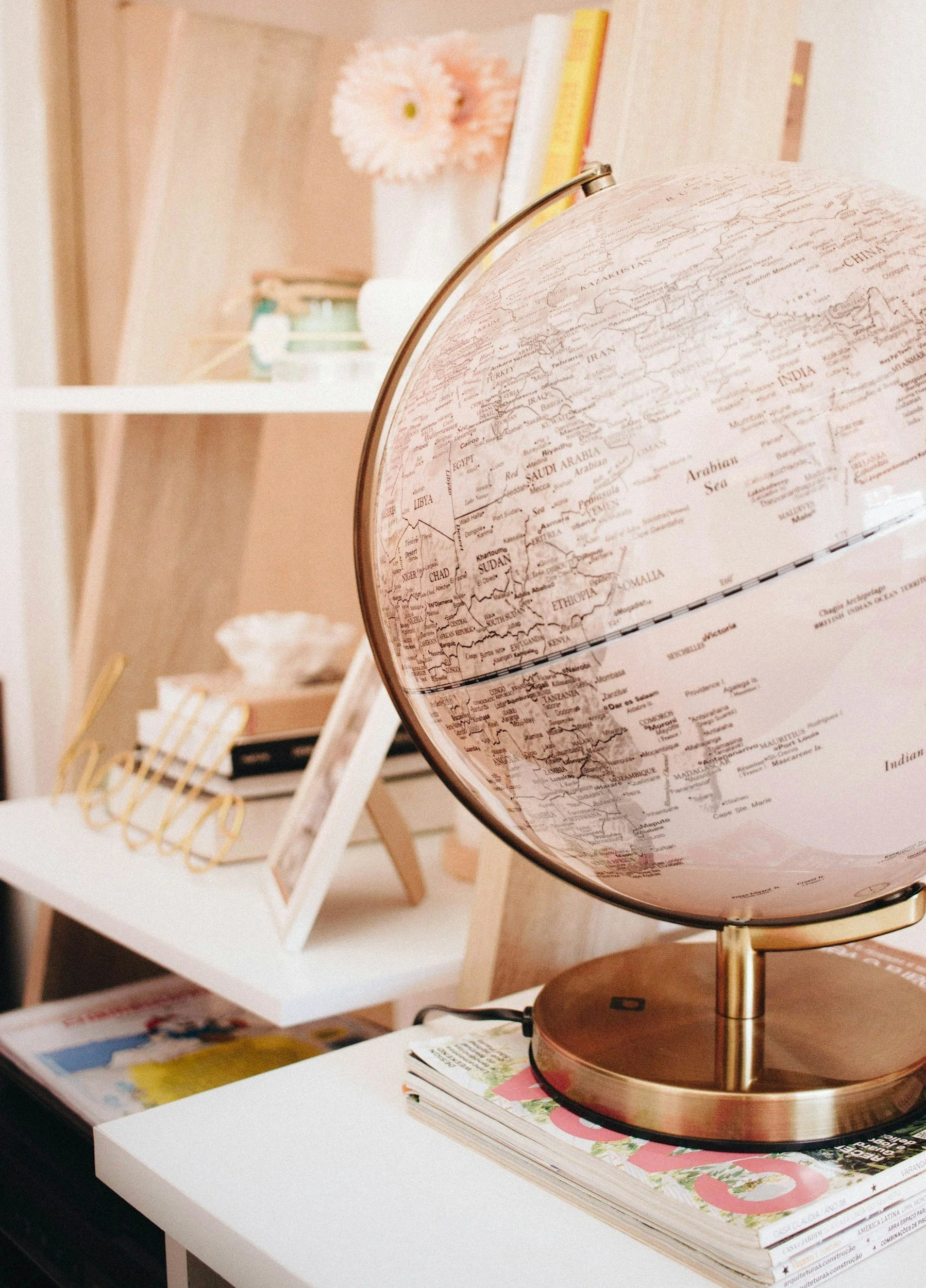 A white table with a stack of magazines and a globe on it. In the background, there is a white bookshelf displaying books, a framed picture, and a small vase with a pink flower.