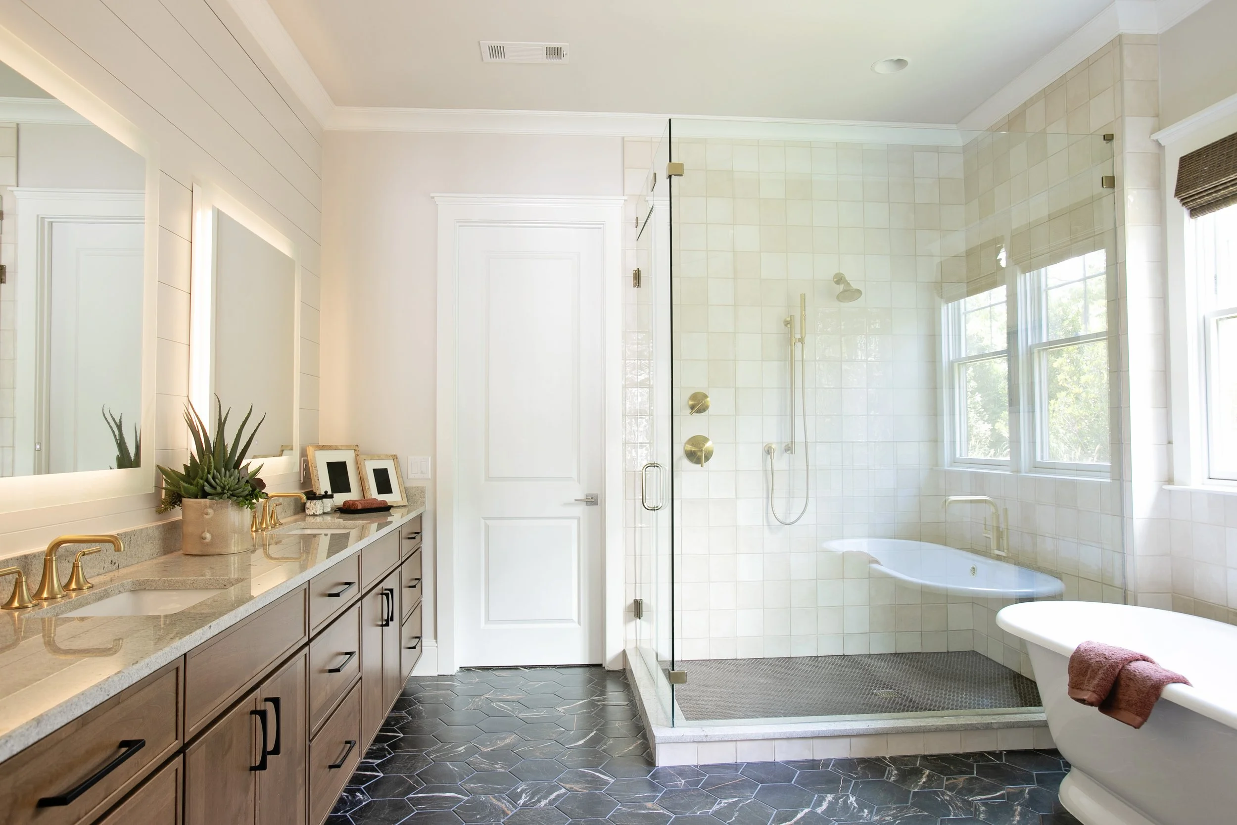 A modern bathroom with a large vanity mirror, potted plant, framed photos, gold fixtures, and a granite countertop. There is a white door, and a glass-walled shower with beige tiles, a showerhead, and large windows letting in natural light. The bathroom has dark hexagonal tile flooring.