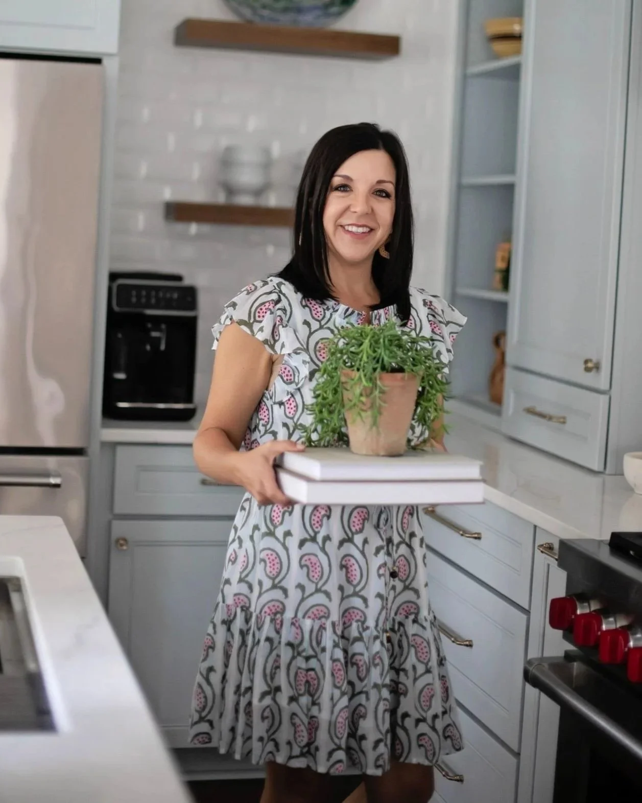 A woman with dark hair smiling in a kitchen, holding a potted succulent plant and two books.