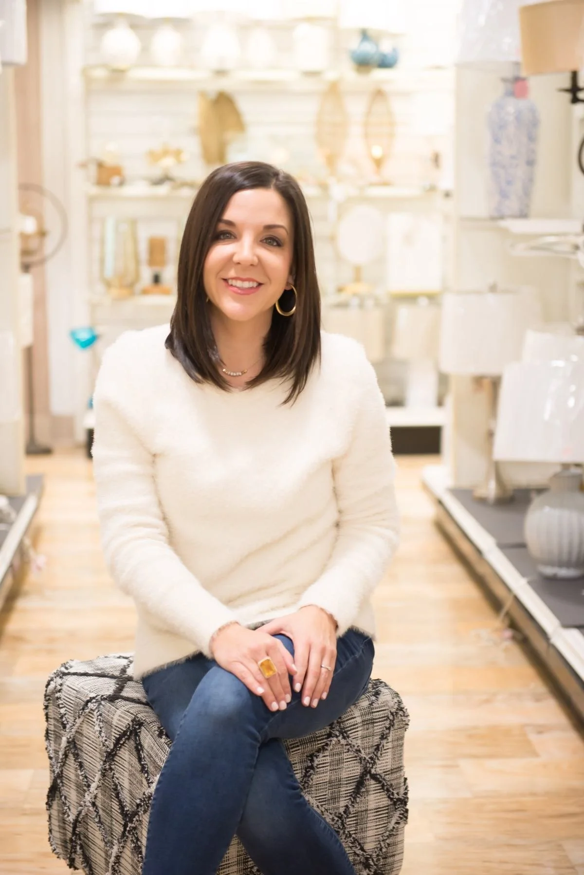 A woman with shoulder-length dark hair, wearing a white sweater, gold hoop earrings, and a gold necklace, sitting on a patterned ottoman in a home decor store. She is smiling, with some jewelry on her fingers and a black cell phone in her hands. The background displays shelves with various vases, bowls, and decorative objects.