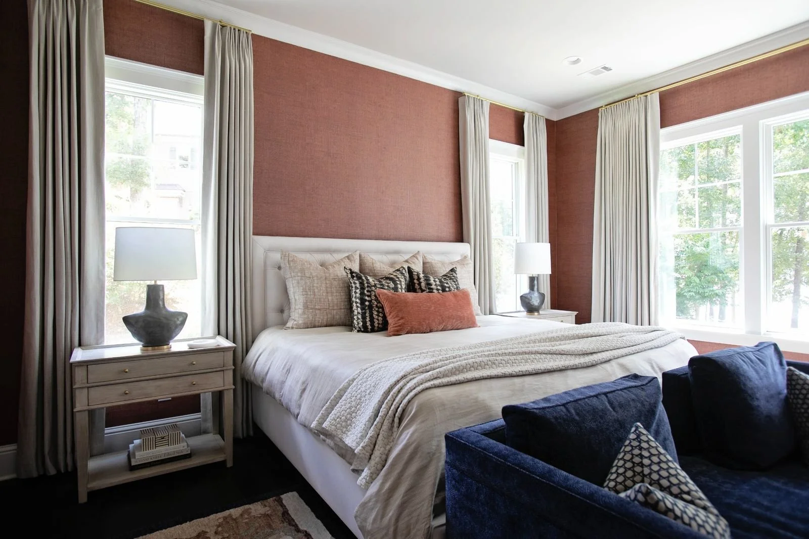 A bedroom with a large bed, beige and patterned pillows, cream-colored bedding, and a blue velvet armchair. There are two nightstands with table lamps, and tall windows with white curtains letting in natural light. The walls are painted a warm reddish-brown.