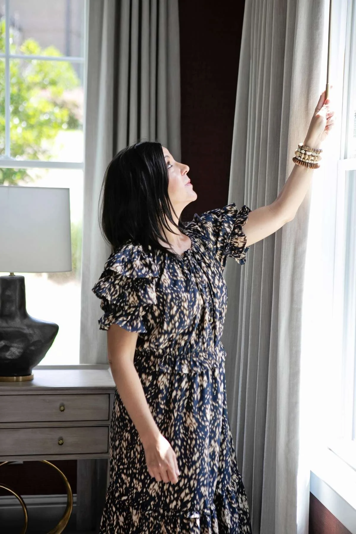 Woman with dark hair wearing a patterned dress, standing by a window, adjusting the curtains.