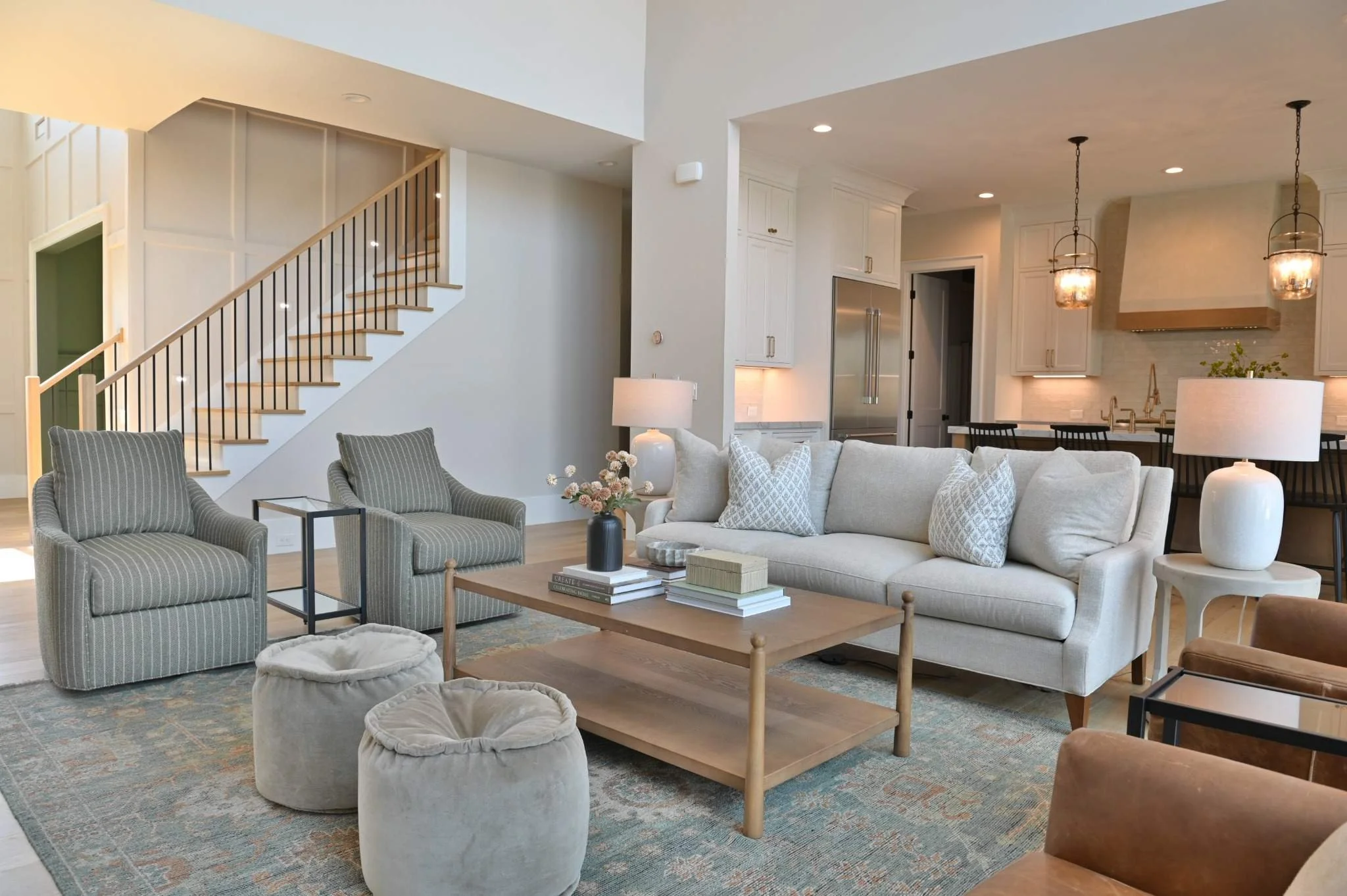 Interior view of a living room with a staircase, a white sofa, armchairs, a coffee table, lamps, and a kitchen area in the background.