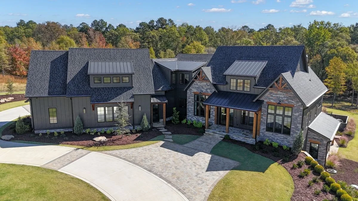 A large modern house with a mix of dark wood siding and gray stone exterior, surrounded by a landscaped yard and winding driveway, with trees in the background.