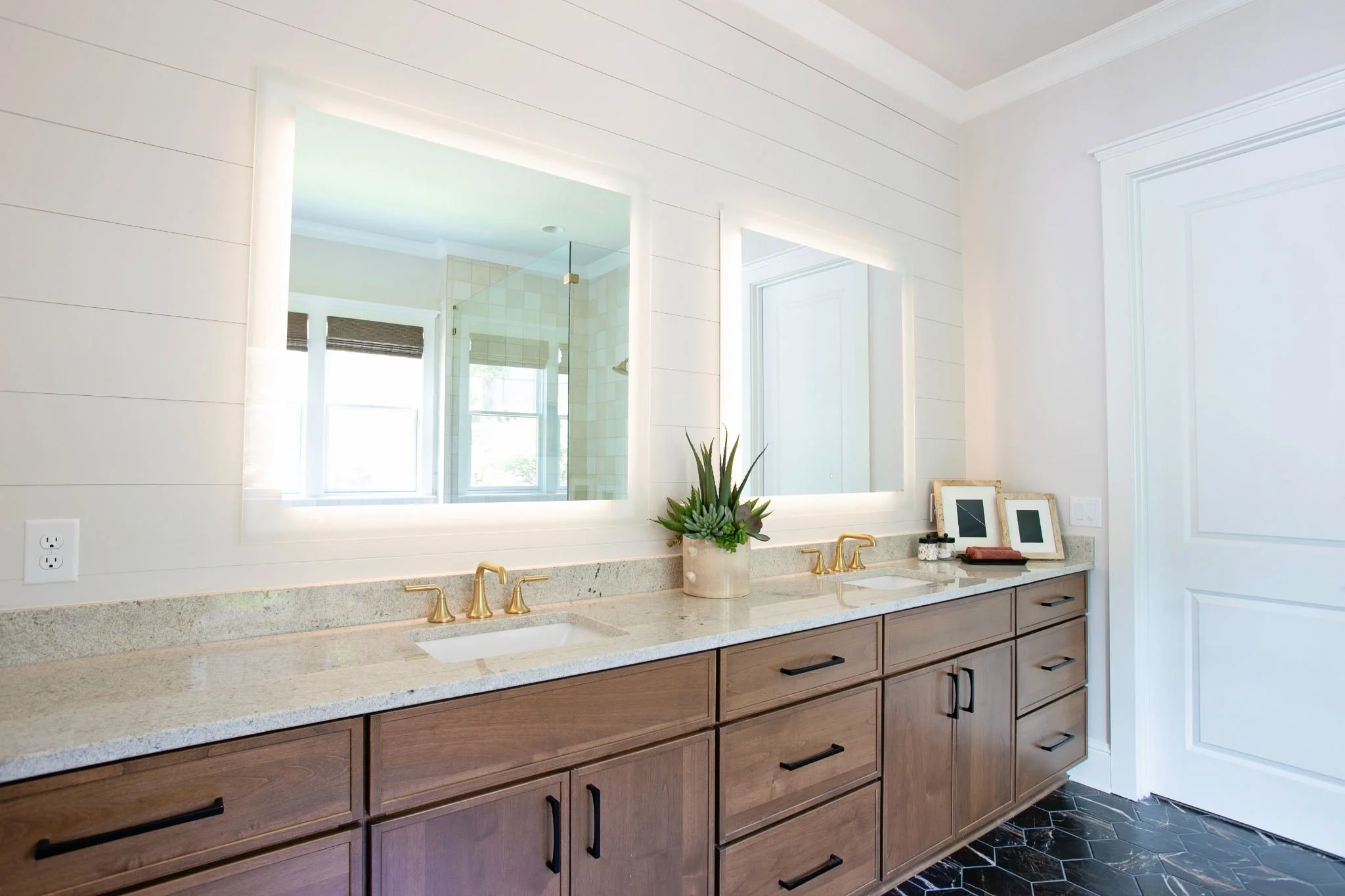 Bathroom with a large mirror, beige countertop with two sinks and gold faucets, wooden cabinet below, decorative plant, framed photos on the counter, black and white patterned tile floor, white walls with trim, and a glass shower enclosure in the background.