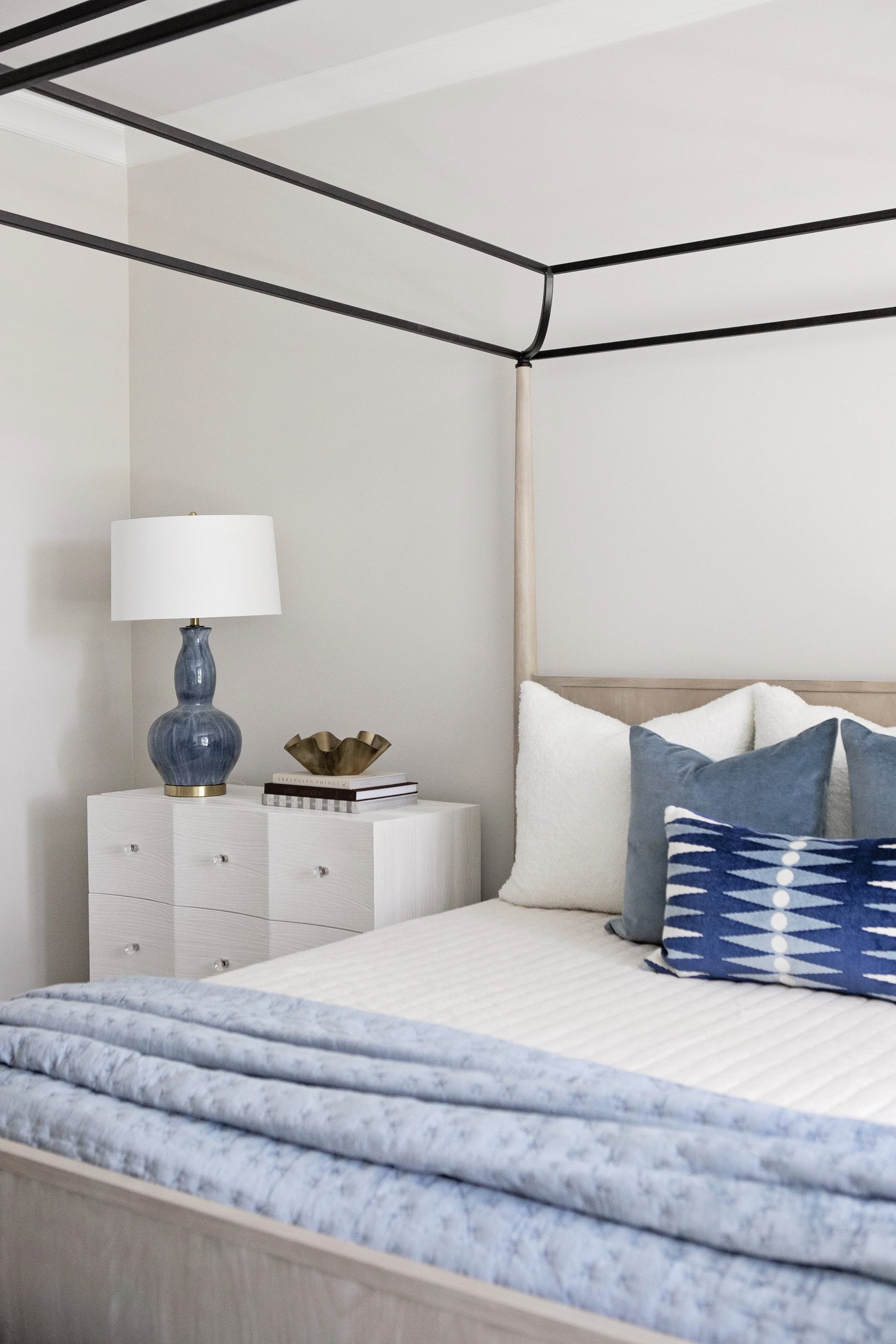 Bedroom with a bed, white side table, blue table lamp, and pillows, with a canopy frame over the bed and a textured blanket.