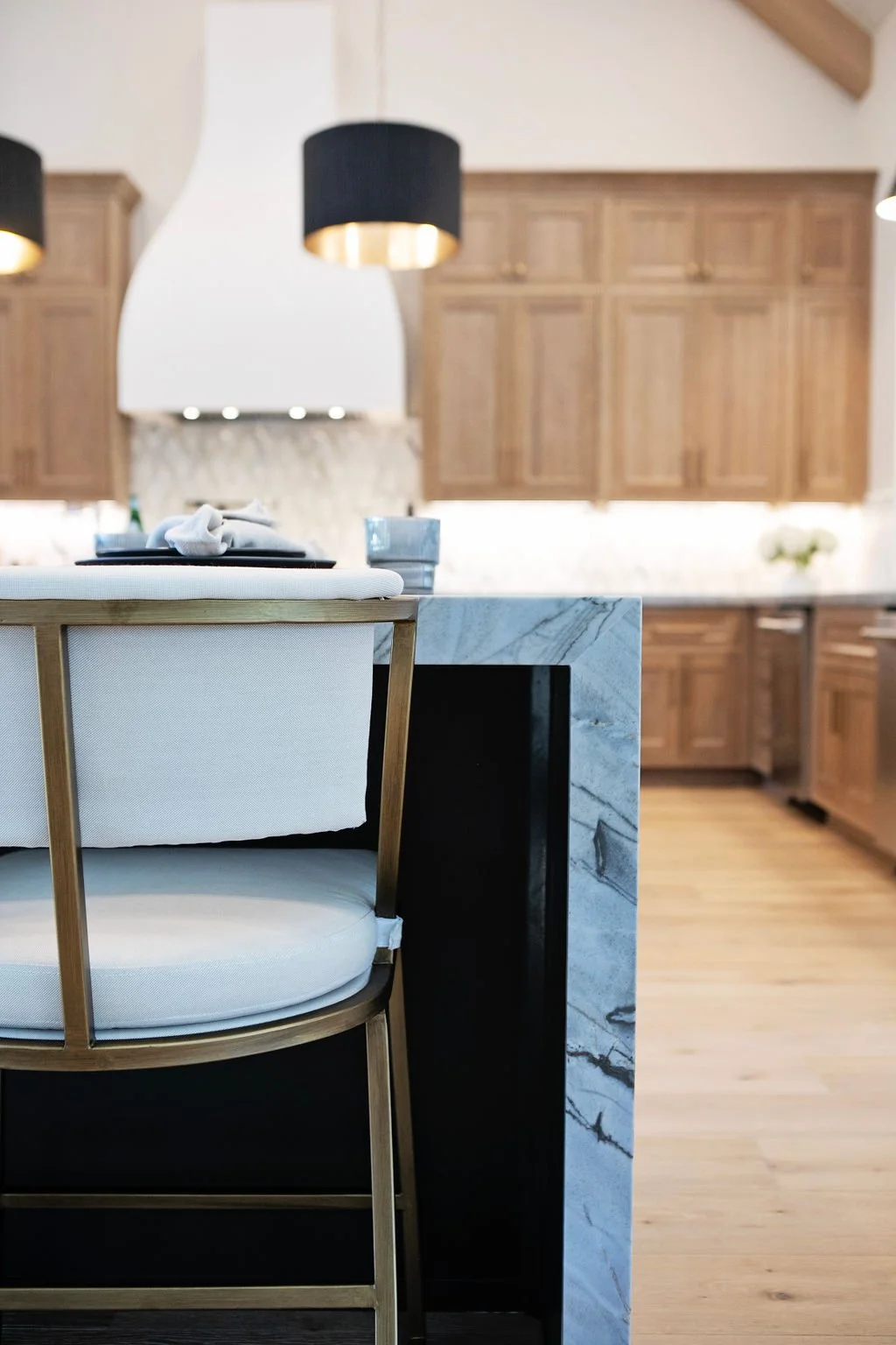 Close-up of a table setting in a modern kitchen, featuring a white placemat, napkin, black plate, and glass on a marble countertop, with wooden cabinets and black pendant lights in the background.