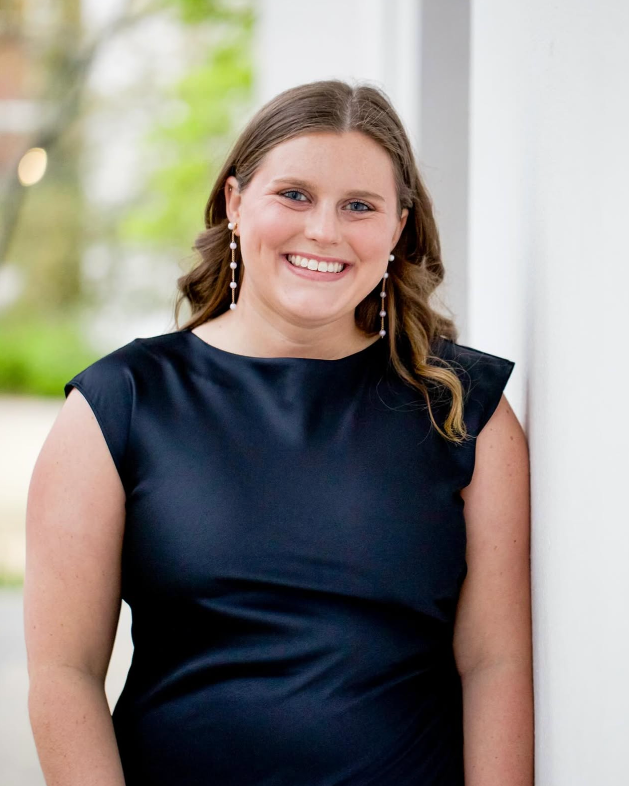 A smiling woman with wavy brown hair, wearing a sleeveless navy blue dress and dangling pearl earrings, standing outdoors beside a white wall with greenery in the background.