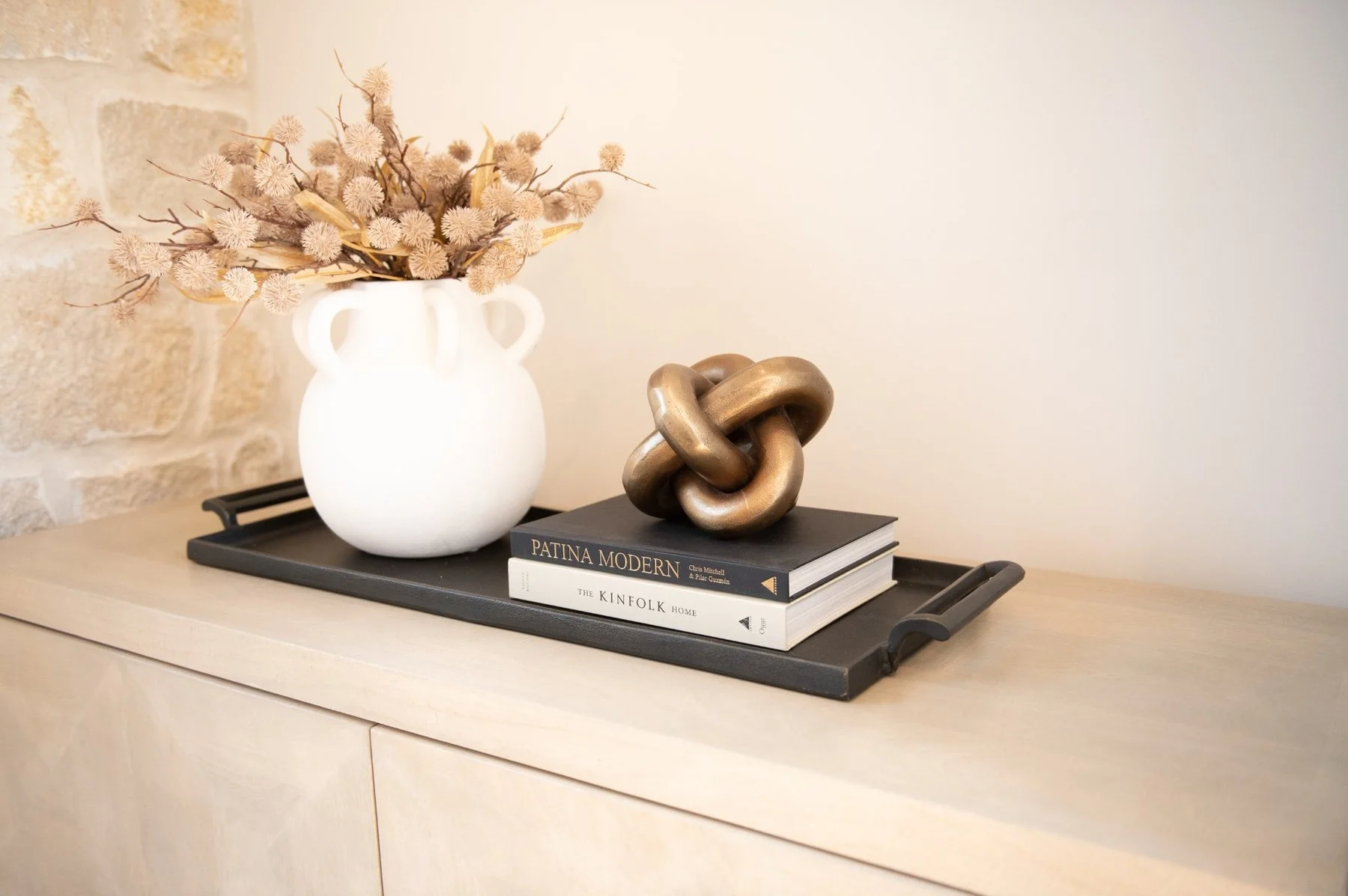 Decorative white vase with dried beige flowers, a metallic knot sculpture, and two coffee table books on a black tray, all placed on a light wood surface against a beige wall.