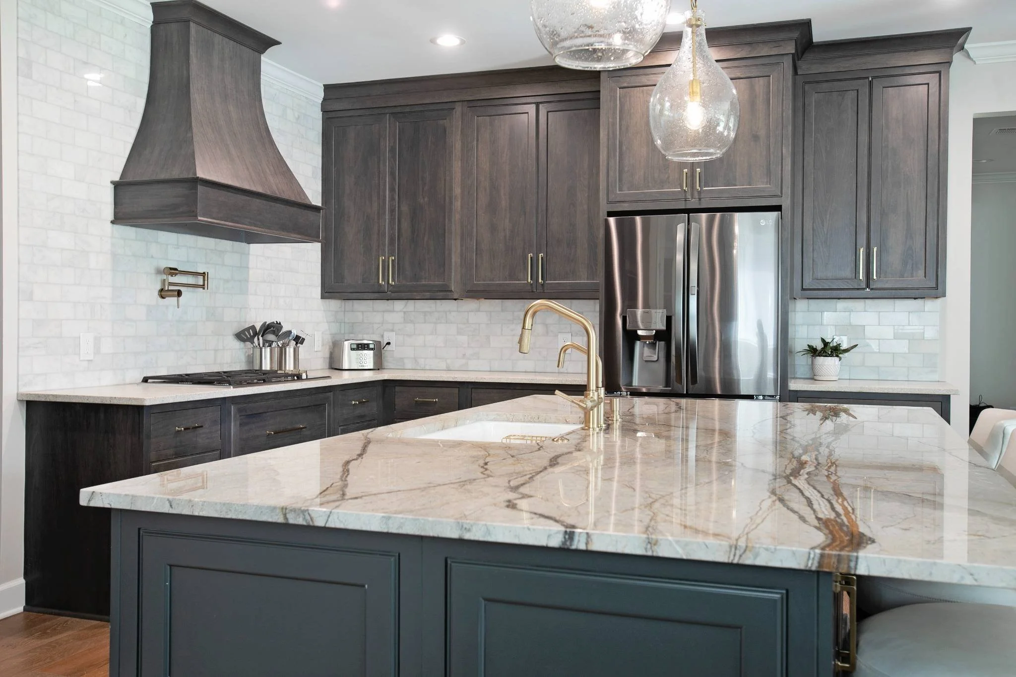 Modern kitchen with dark wood cabinets, a white marble island, stainless steel refrigerator, white subway tile backsplash, gold faucet, and two glass pendant lights.