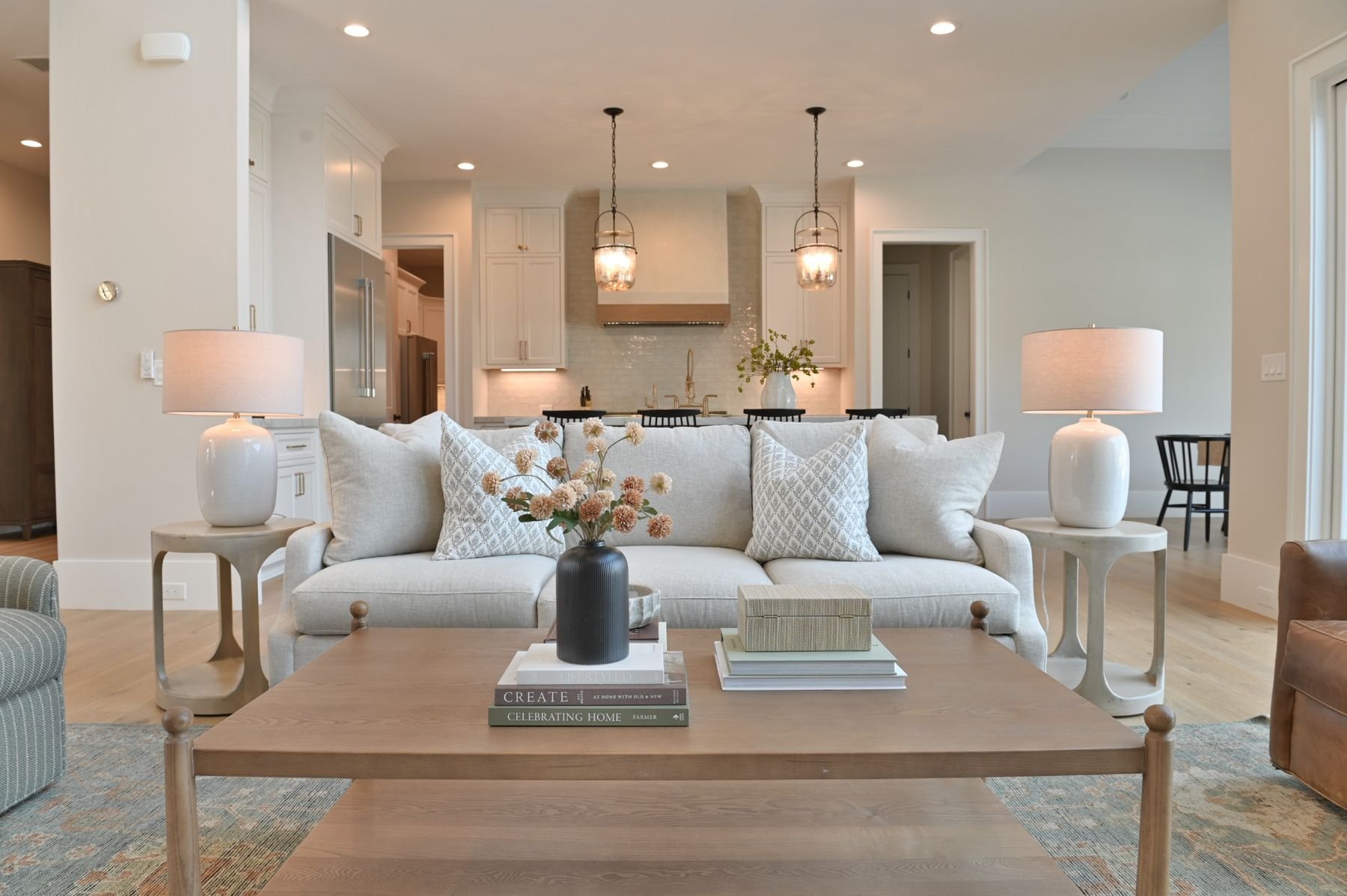 Living room with a cream sofa, decorative pillows, and a wooden coffee table with books and a vase of flowers. Two side tables with white lamps flank the sofa. The background shows a kitchen with white cabinets, pendant lights, and a vase with greenery on the kitchen counter.