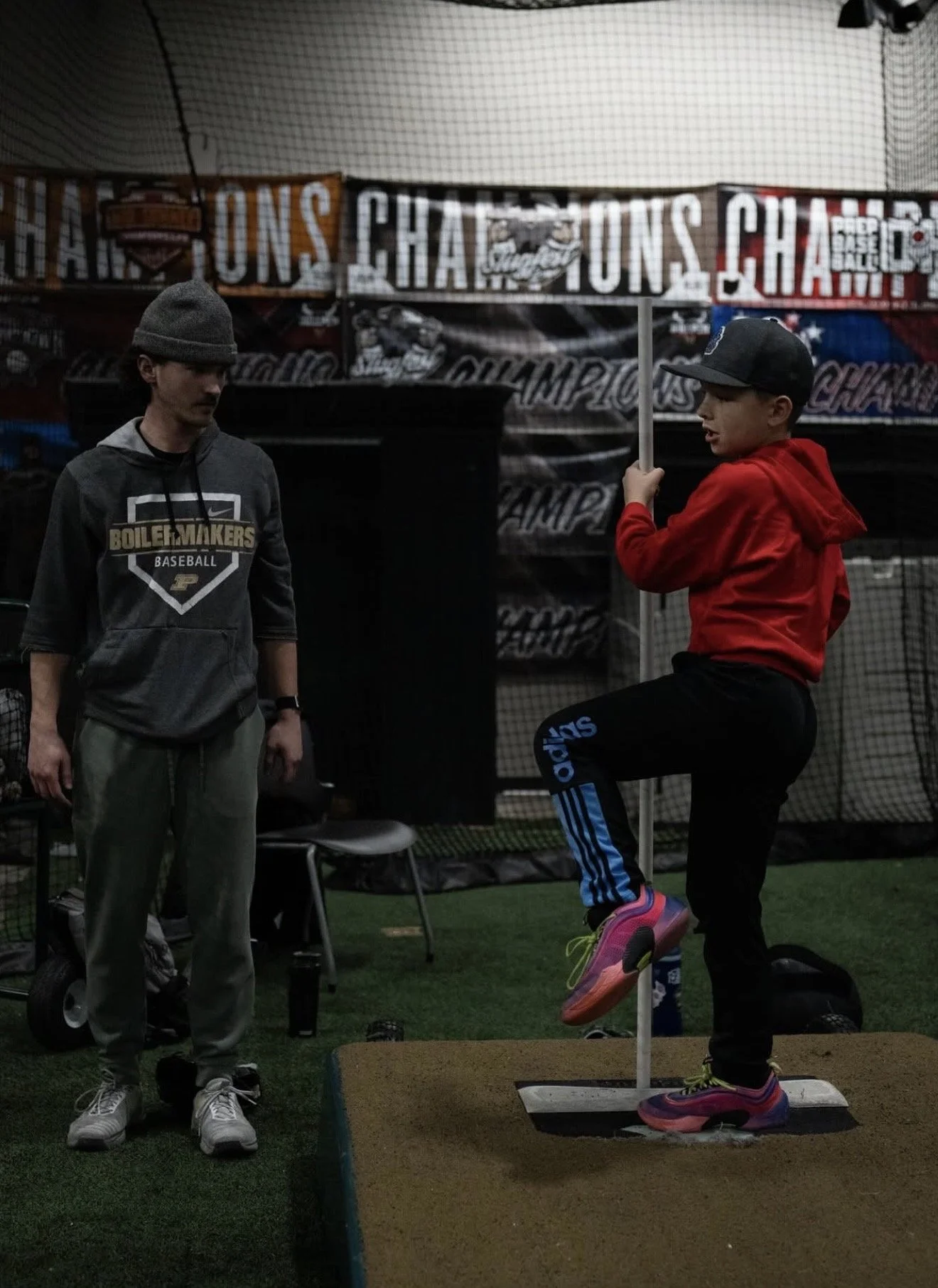 A young girl in a red hoodie and colorful sneakers is practicing a long jump at an indoor sports facility. A man, possibly a coach or trainer, stands nearby watching her. The background features banners with the word 'CHAMPIONS' and sports-themed images.