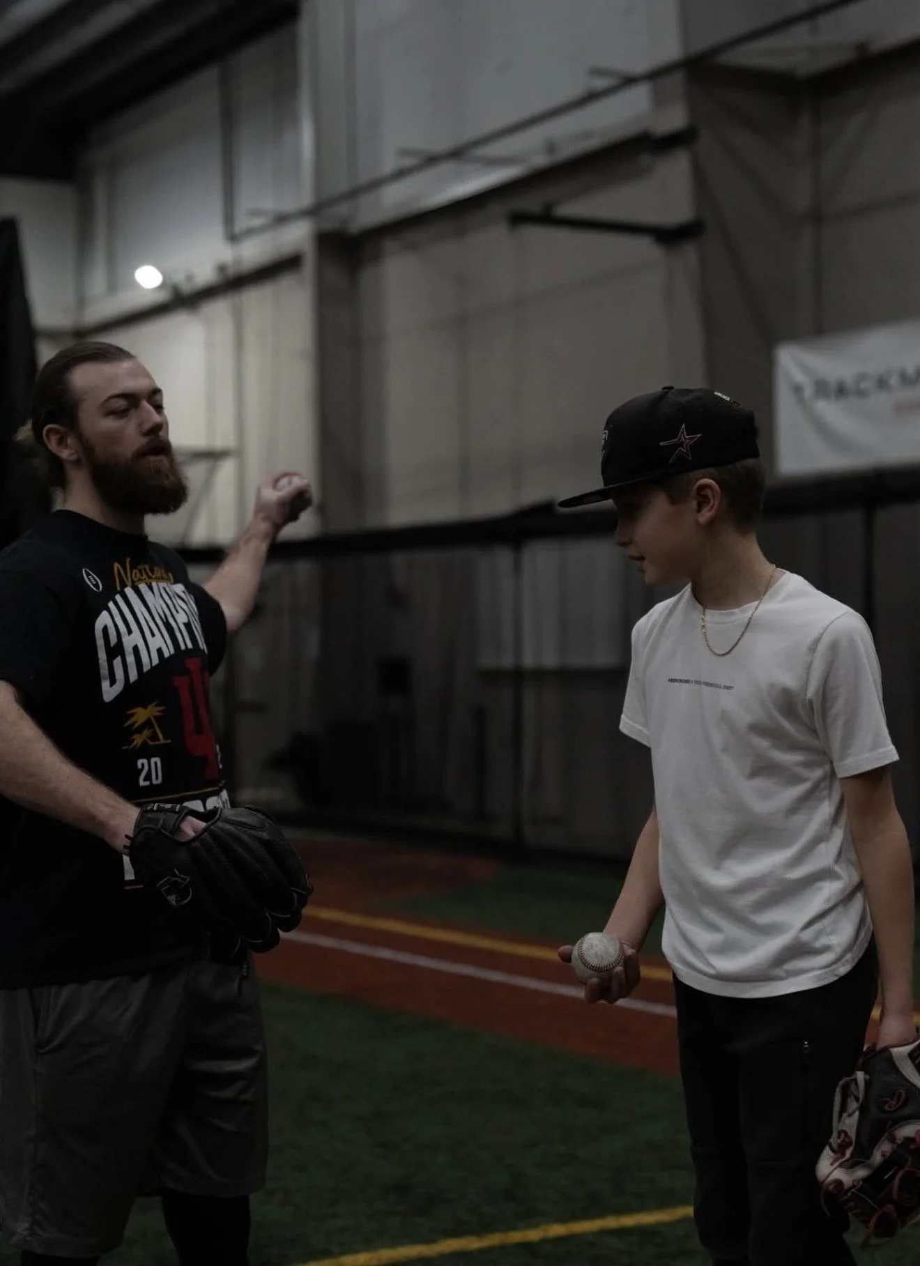 Two males, one with a beard wearing a black T-shirt and baseball glove, and the other with short hair and a baseball cap, talking inside a sports facility with a batting cage net in the background. The young man is holding a baseball in his hand.