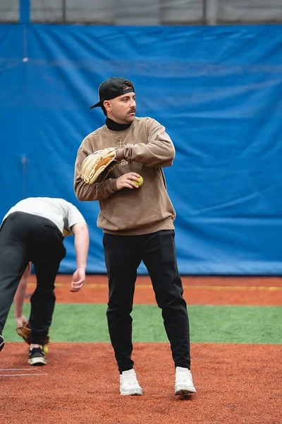 A young man wearing a black cap, brown sweatshirt, and black pants standing on a baseball field, holding a baseball glove and ball, with a blue wall in the background.