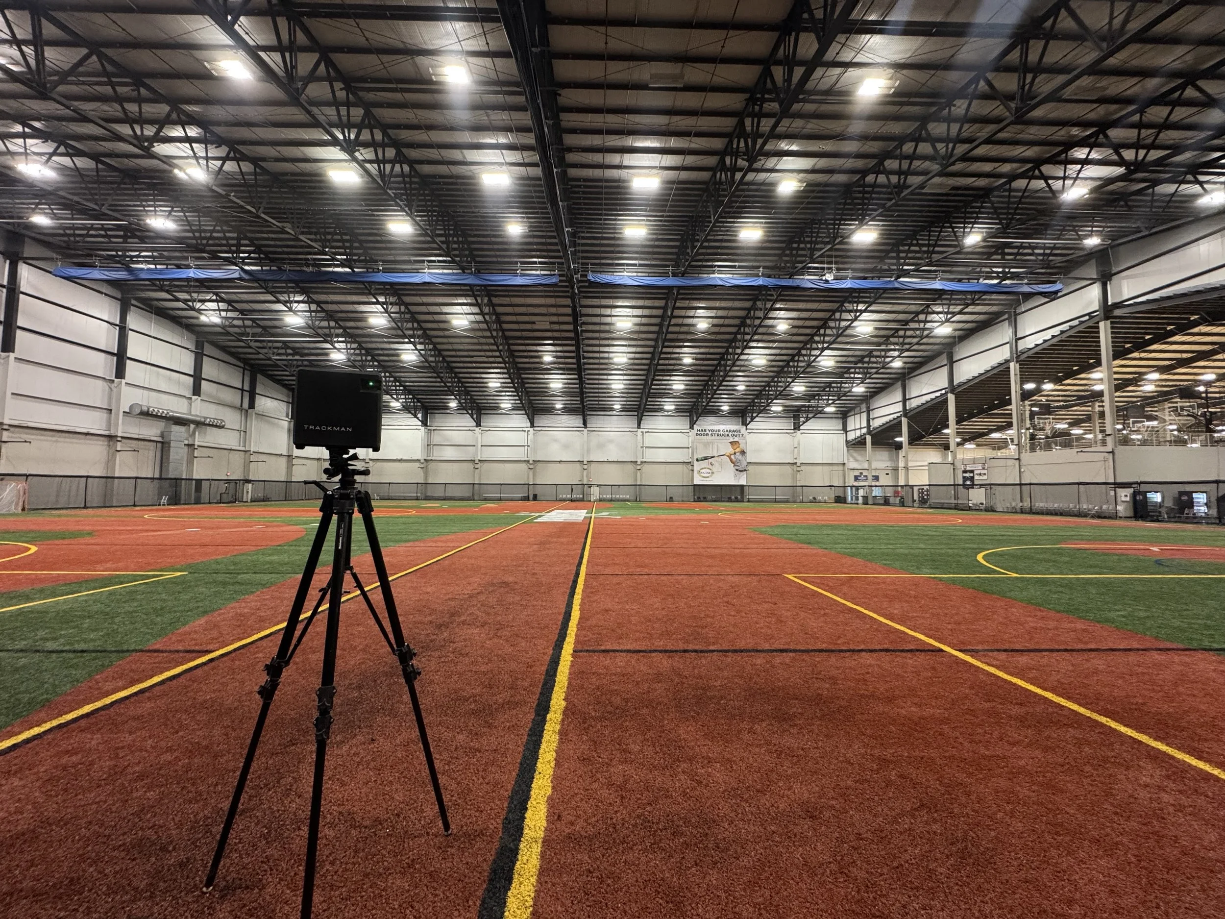 Indoor sports facility with a large open field, high ceiling, and bright overhead lights. There is a tripod with a camera or projector in the foreground and yellow and red lines on the floor for sports markings. A sign on the wall reads ‘Has Your Garage Door Struck Out?’