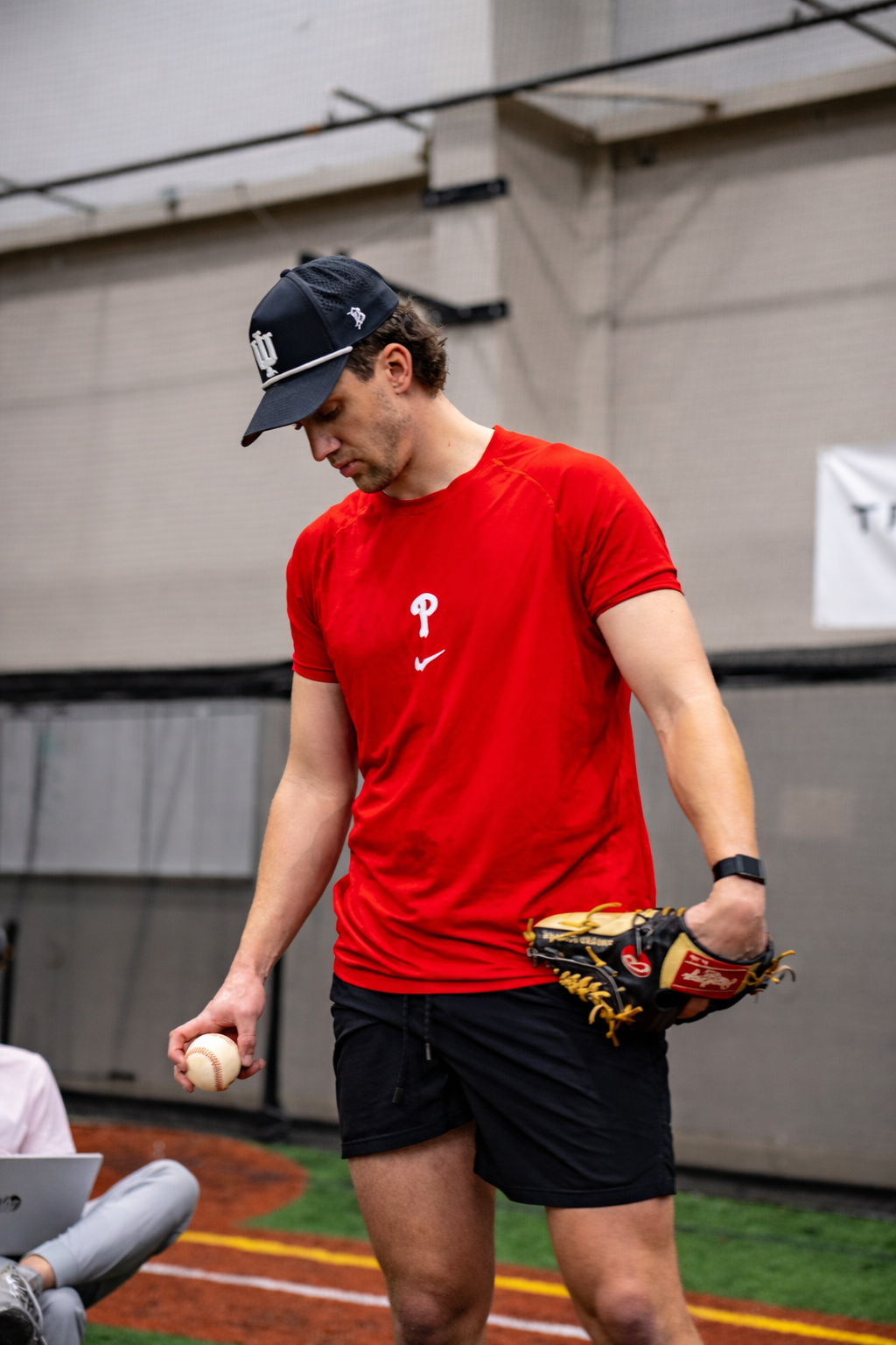 A man in a red t-shirt and black shorts is holding a baseball in one hand and a baseball glove in the other, inside an indoor sports facility.