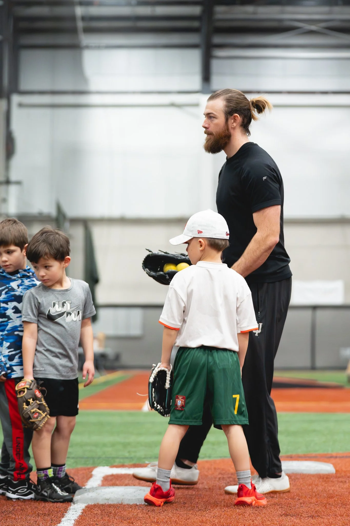A young male baseball coach with a beard, tied-back hair, wearing a black shirt, is instructing a group of young children on a baseball field indoors. The children are wearing baseball gear, including a white cap, a gray Nike shirt, and green shorts. The coach is holding a baseball glove and multiple yellow baseballs. The children are standing near a white home plate on the artificial turf field.