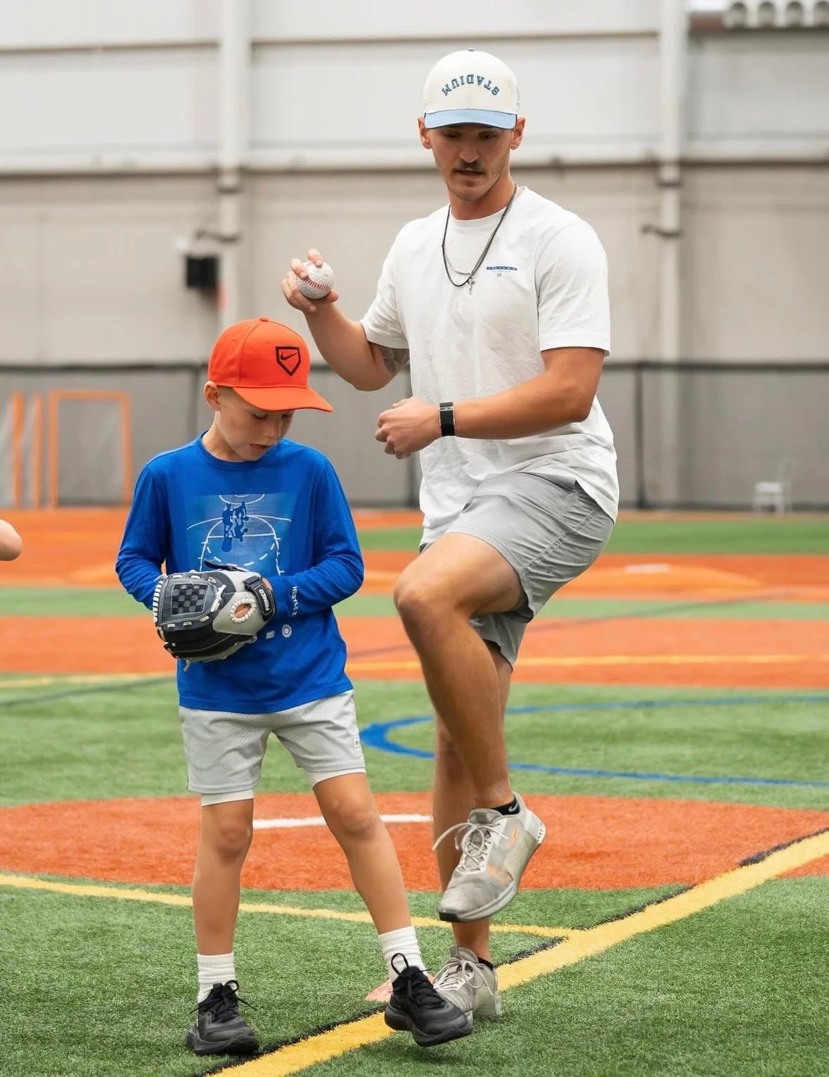 A man coaching a young boy on baseball in an indoor sports facility. The man holds a baseball and is demonstrating a stance, while the boy wears a baseball glove and cap, looking at the ground.