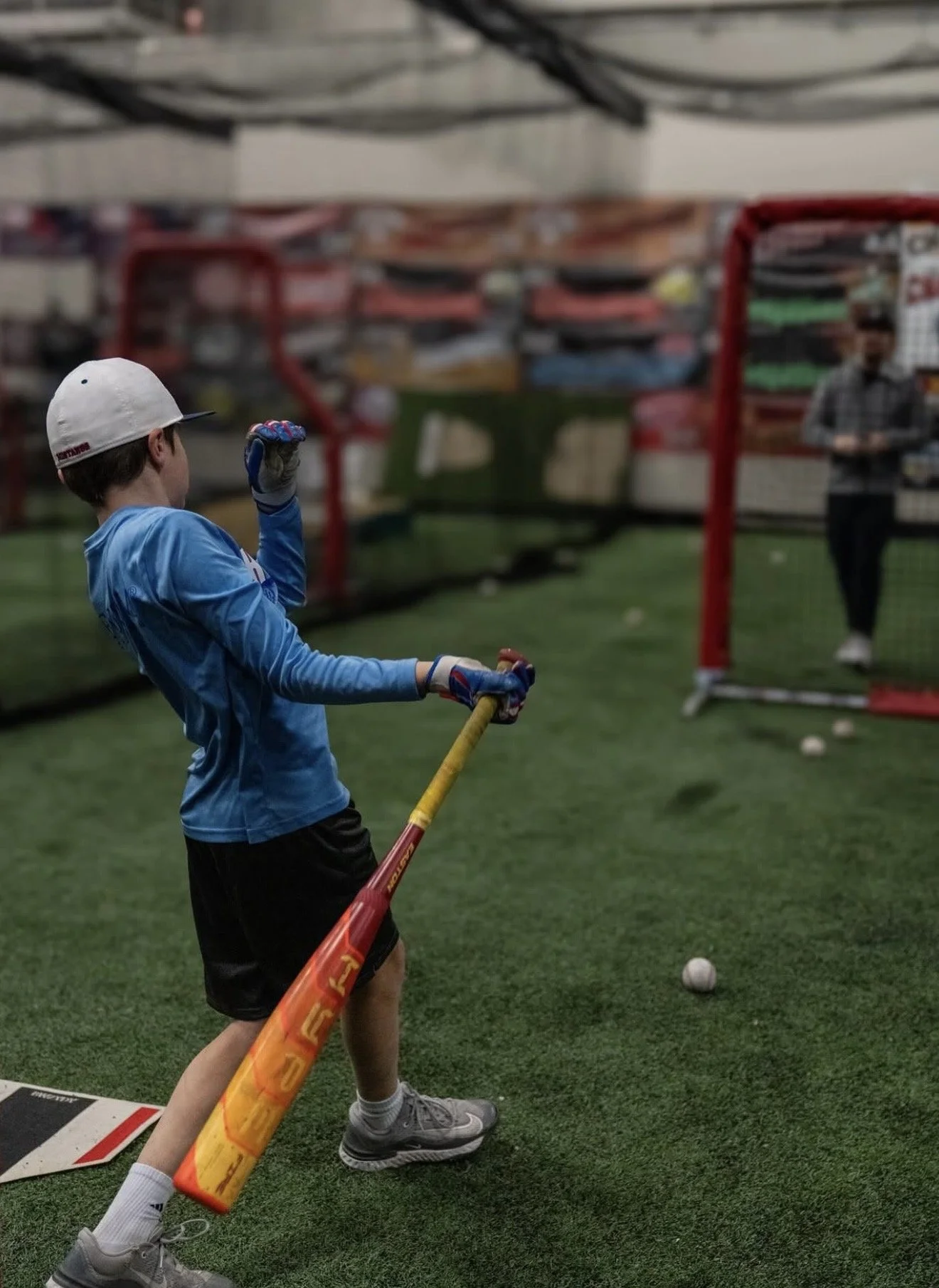 A young boy in a blue jersey and white helmet practicing baseball with a yellow bat in an indoor batting cage. An adult man stands in the background near the net, watching him.