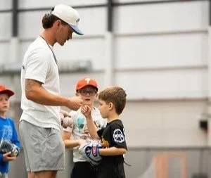 Young coach handing a medal to a young boy during a youth sports event, with other children and a sports facility in the background.