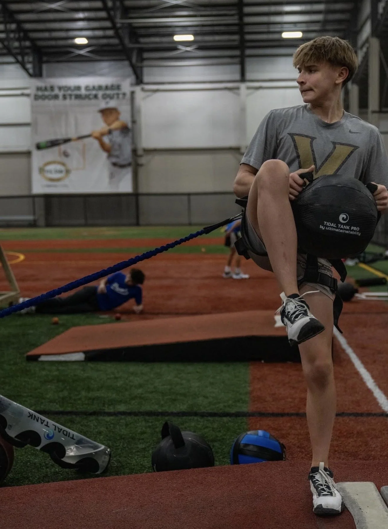 A young male athlete stretching his leg on a knee elevation foam pad during indoor training. He is wearing a gray t-shirt with a gold letter 'V' and Nike logo, and black and white running shoes. In the background, another person is seen lying on the ground and a third person is holding a baseball bat in front of a large banner with a young player. The training area has red and green turf and various fitness equipment.