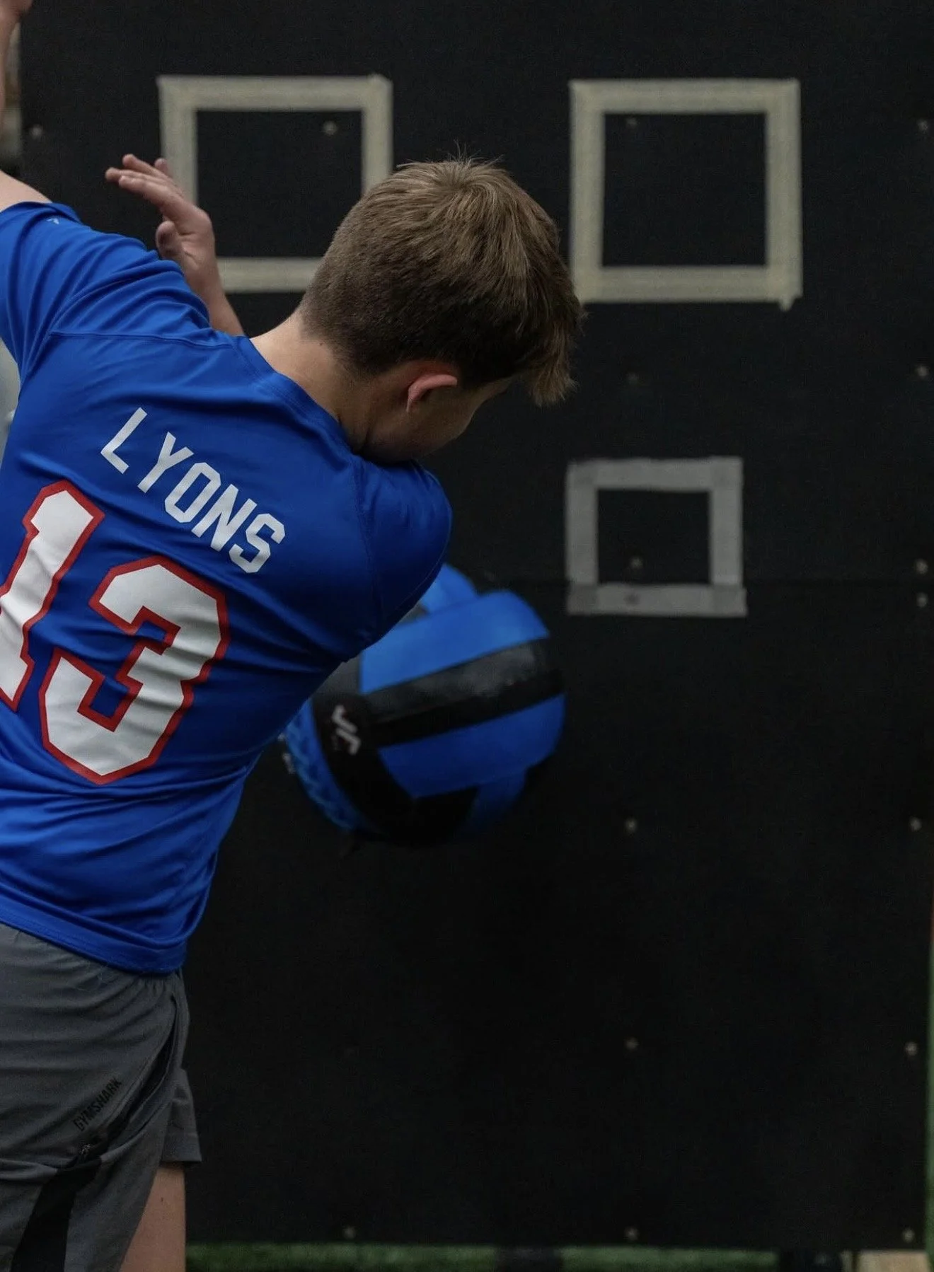 A man in a blue sports jersey with the name 'LYONS' and the number 13 is throwing a football, standing indoors against a black wall with two white rectangles.