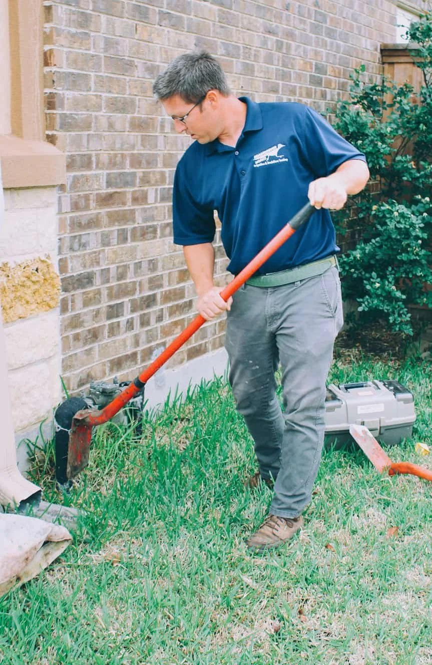 A man in a blue polo shirt and gray pants using an orange hoe to work on the ground next to a brick wall, with a toolbox nearby.