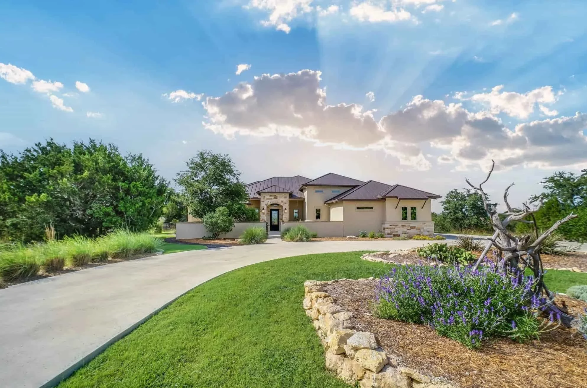 Wide view of a modern house with a landscaped front yard, green grass, flower beds, and a curved driveway under a partly cloudy sky.