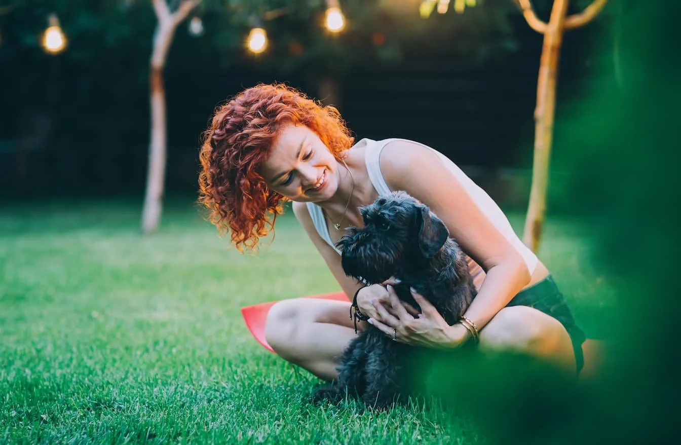 A woman with curly red hair, wearing a white tank top and black shorts, sitting on grass and smiling while holding a small black dog outdoors in the evening.