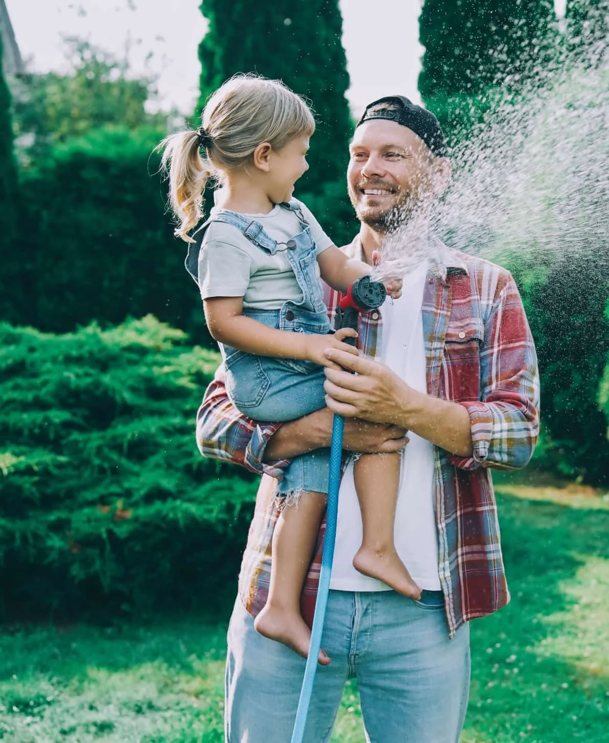 A father and daughter enjoying playtime outside, with the father holding the daughter and the daughter spraying water from a hose at her father, both smiling in a garden.