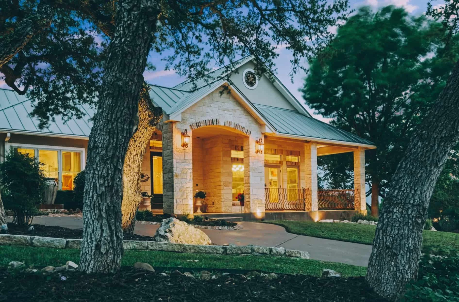 A charming house with a stone exterior and metal roof, illuminated by outdoor lights, surrounded by trees and a well-maintained lawn at dusk.