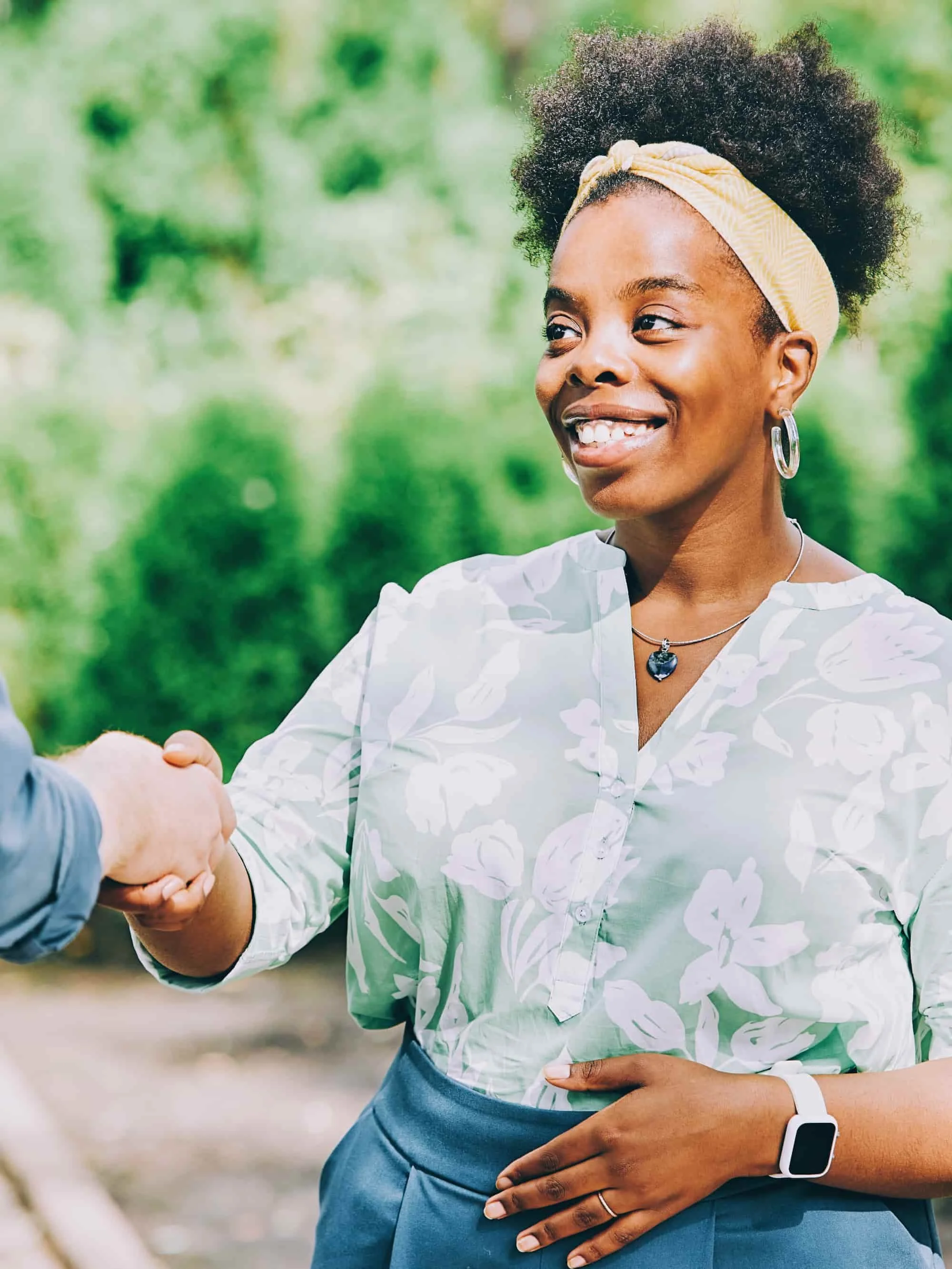 A woman with a natural curly hairstyle wearing a yellow headband, jewelry, a light floral blouse, and a smartwatch shakes hands with someone outside in a green park.