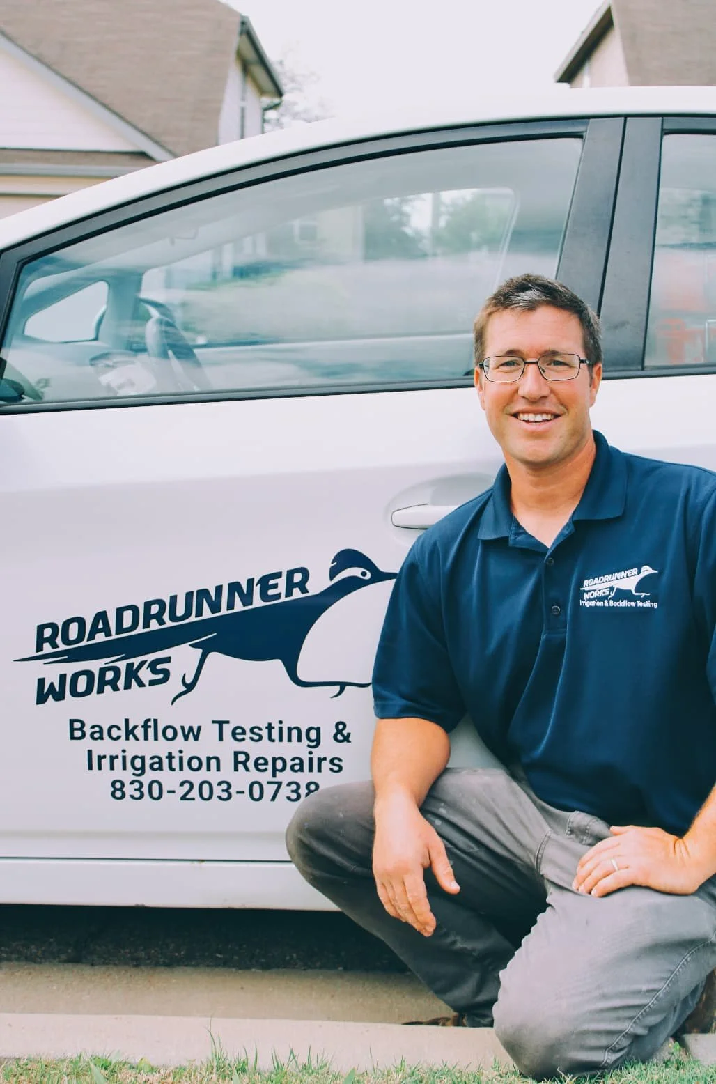 A man with glasses smiling while kneeling in front of a service vehicle with a logo and contact information for Roadrunner Works, a backflow testing and irrigation repair company.